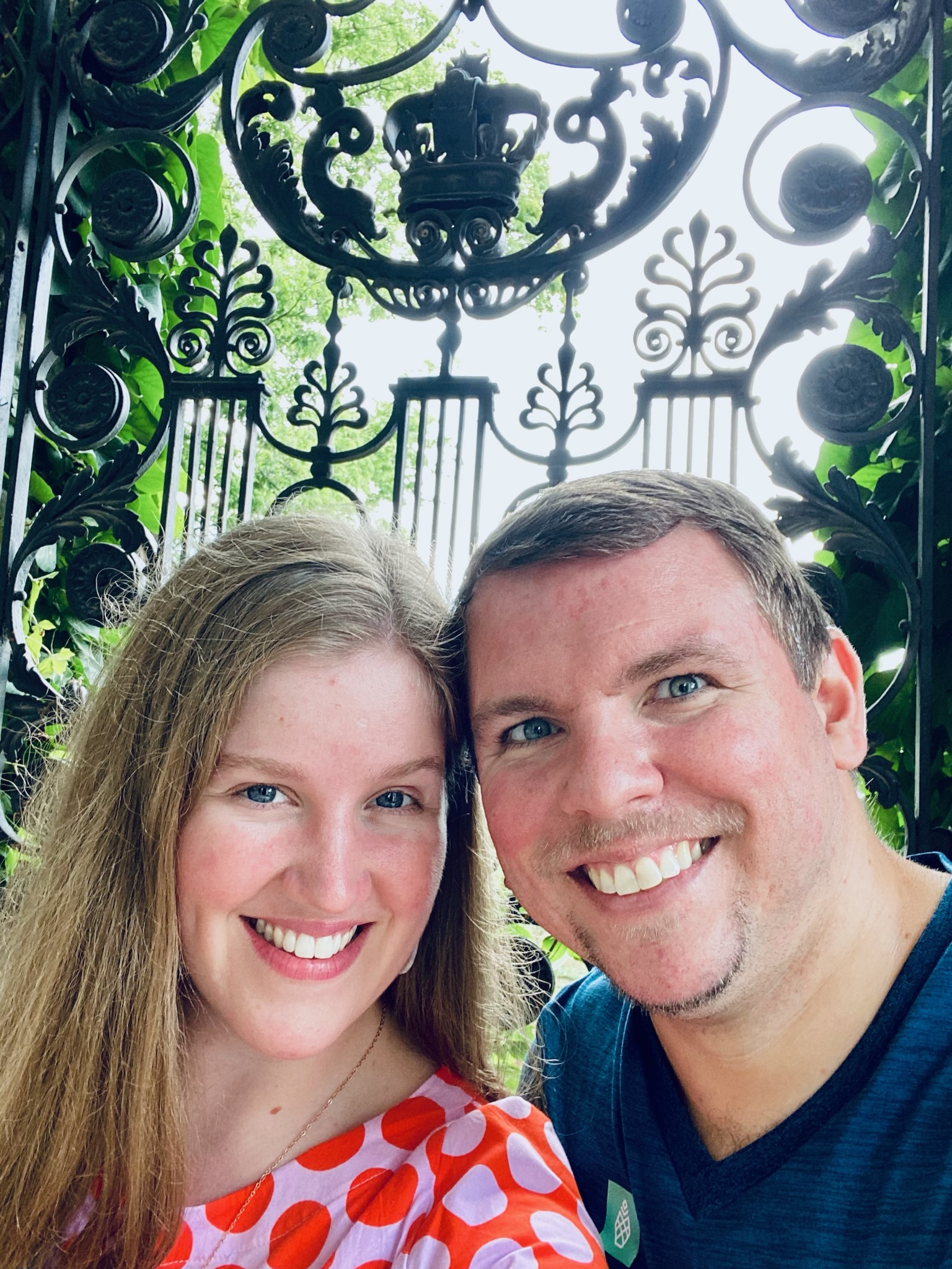 A smiling man and woman take a selfie in front of an ornate black wrought iron gate with intricate designs and greenery in the background.