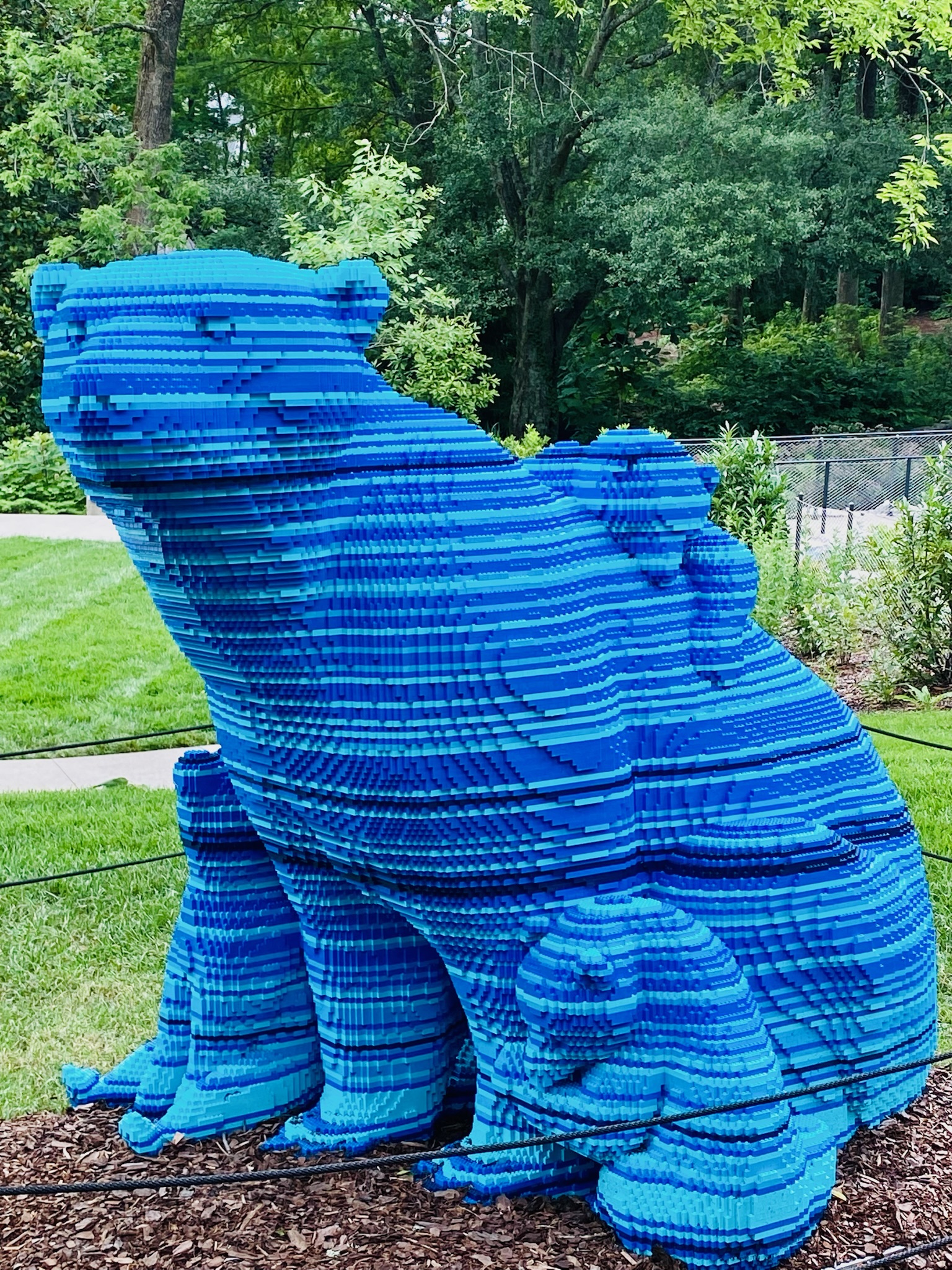 A large blue bear sculpture made of stacked LEGO bricks, with two smaller bear cubs beside it, displayed outdoors on a mulched area near grass and trees.