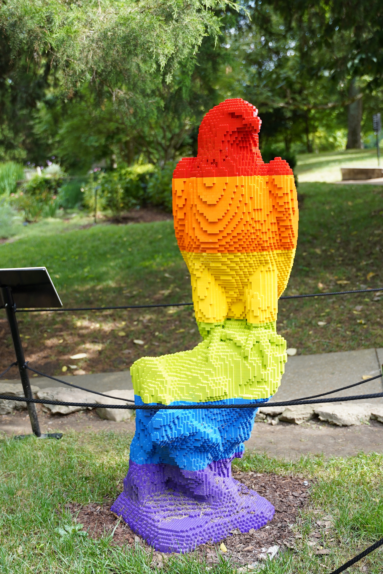 A colorful parrot sculpture made from LEGO bricks sits on a pedestal outdoors, with grass and trees in the background.