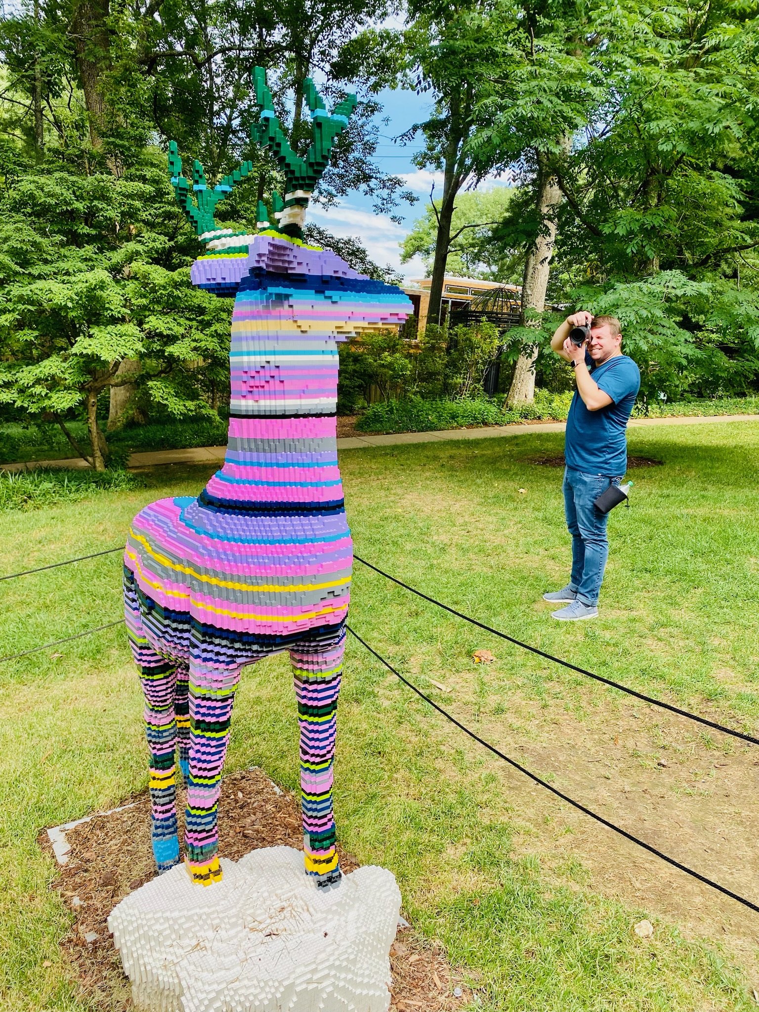 A man takes a photo of a large, colorful deer sculpture made from LEGO bricks in an outdoor park setting surrounded by trees.