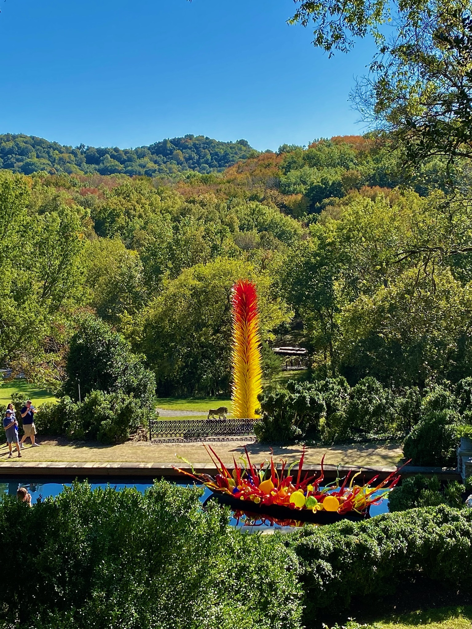 Outdoor garden scene in Nashville with lush green trees, a tall red and yellow glass sculpture in the background, and a rounded glass sculpture with spikes near a reflective pond.