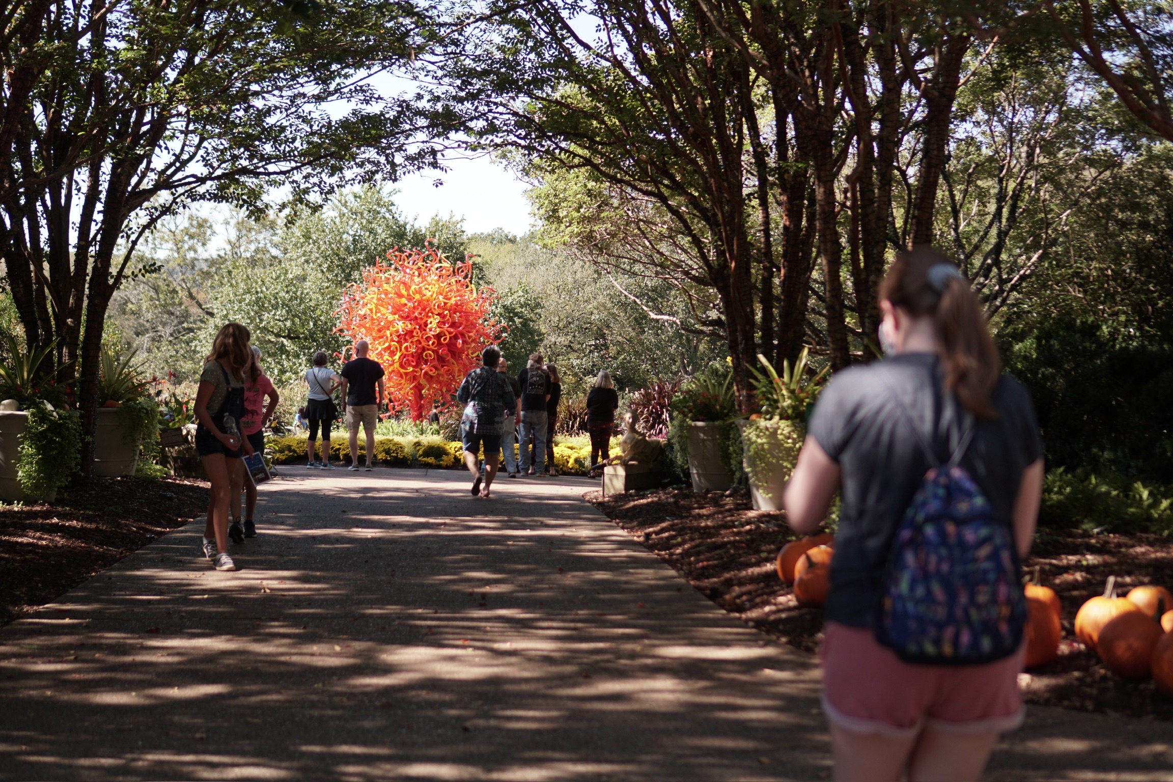 People walk along a tree-lined path toward a bright orange glass sculpture in a Nashville garden on a sunny day.