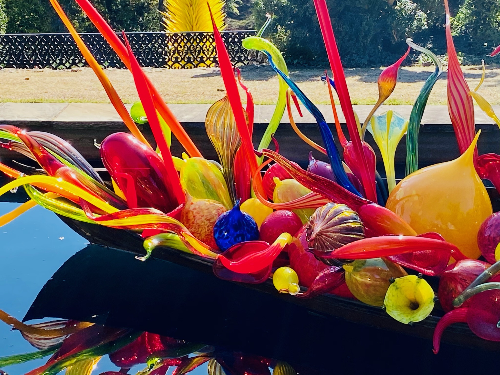 Colorful glass sculptures in various shapes and sizes are arranged in a wooden boat, reflected in the water below, with greenery in the background—an artistic scene reminiscent of Nashville’s vibrant creative spirit.