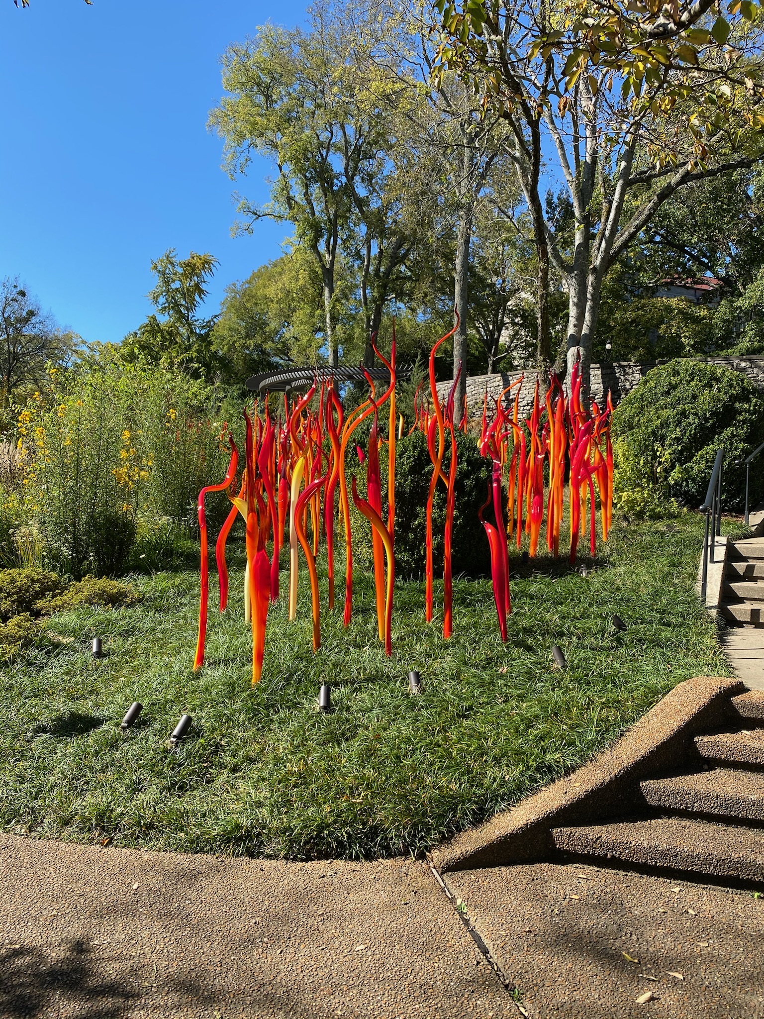 Tall, thin orange and red glass sculptures are arranged in a grassy outdoor garden area with trees, plants, and a stone staircase nearby in Nashville.