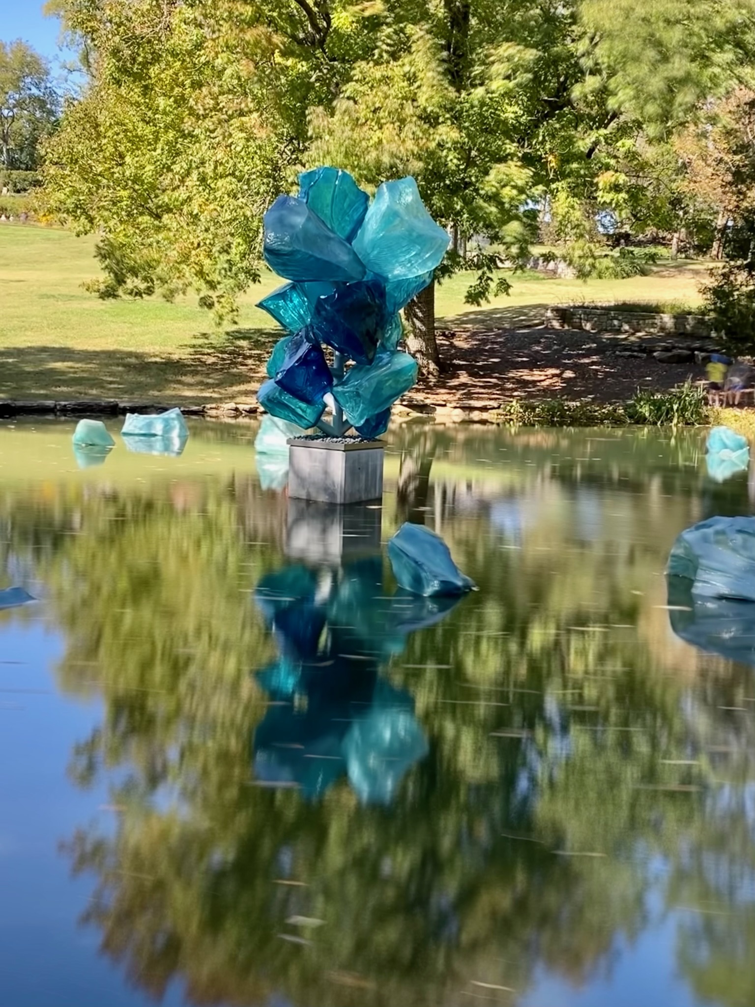 A blue abstract sculpture stands on a pedestal in a pond in Nashville, surrounded by smaller blue shapes, with trees and grass in the background.