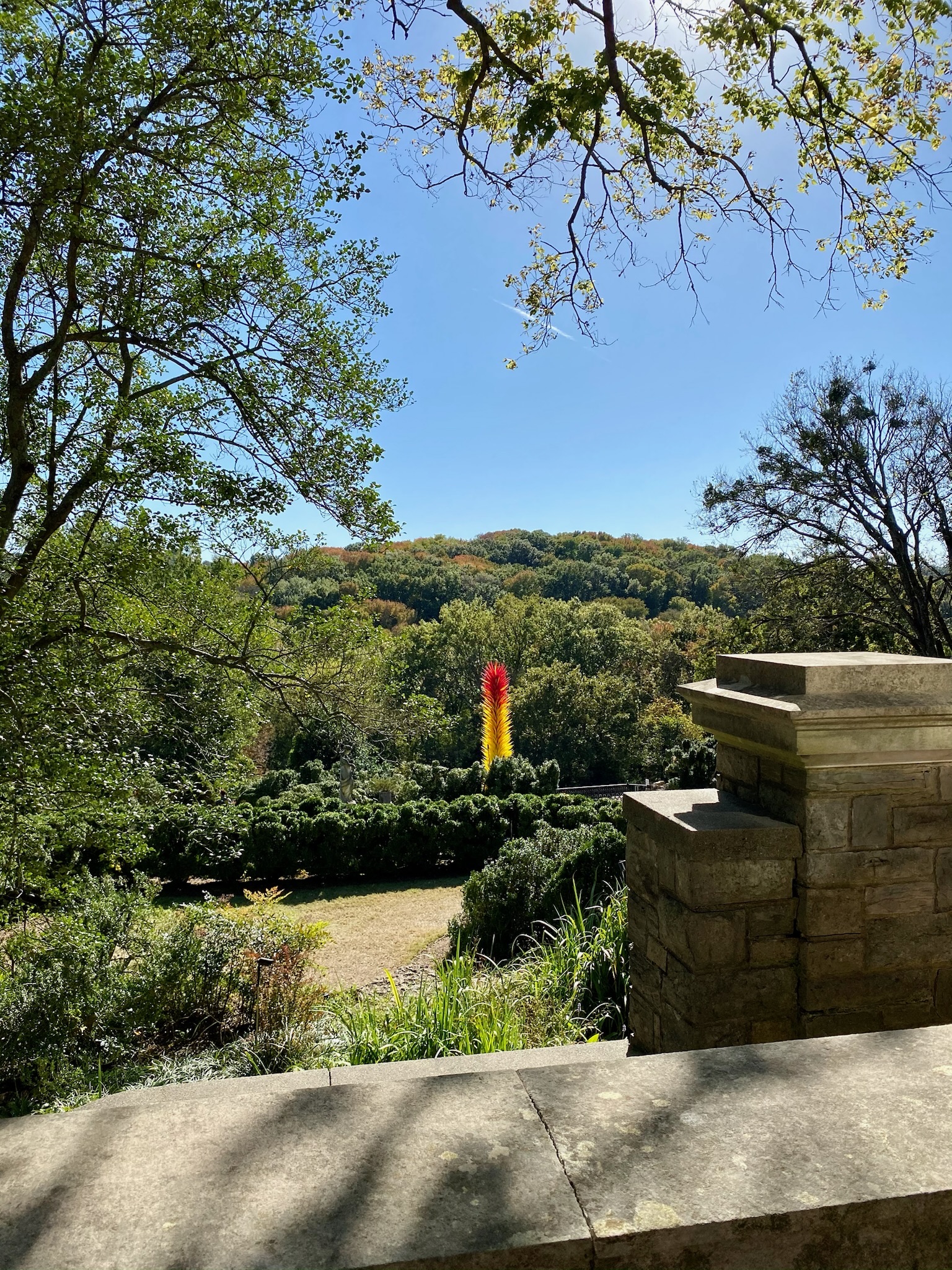 View of a lush green Nashville landscape with a yellow and red vertical sculpture in the center, partially framed by stonework in the foreground and tree branches overhead.