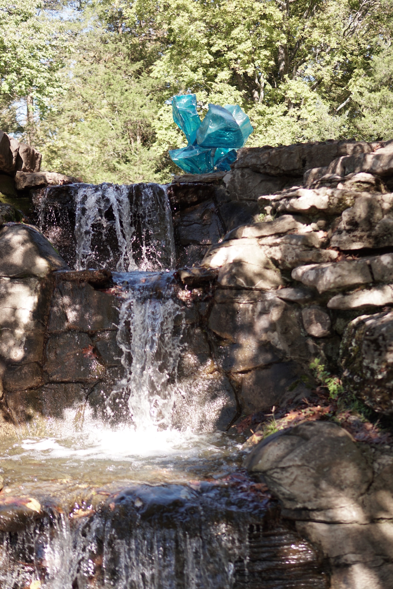 A small waterfall flows over rocks in a wooded Nashville area, with a bright blue abstract sculpture placed near the top of the falls.