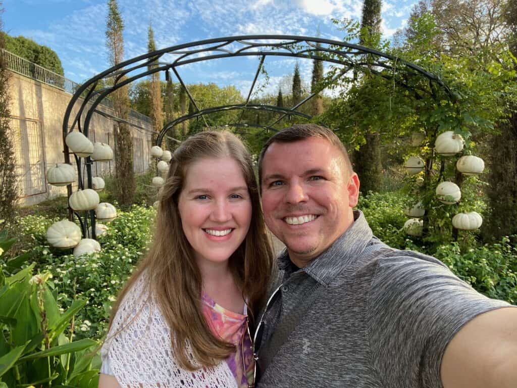 A man and woman smile for a selfie in a garden with a metal arch and hanging white lanterns.