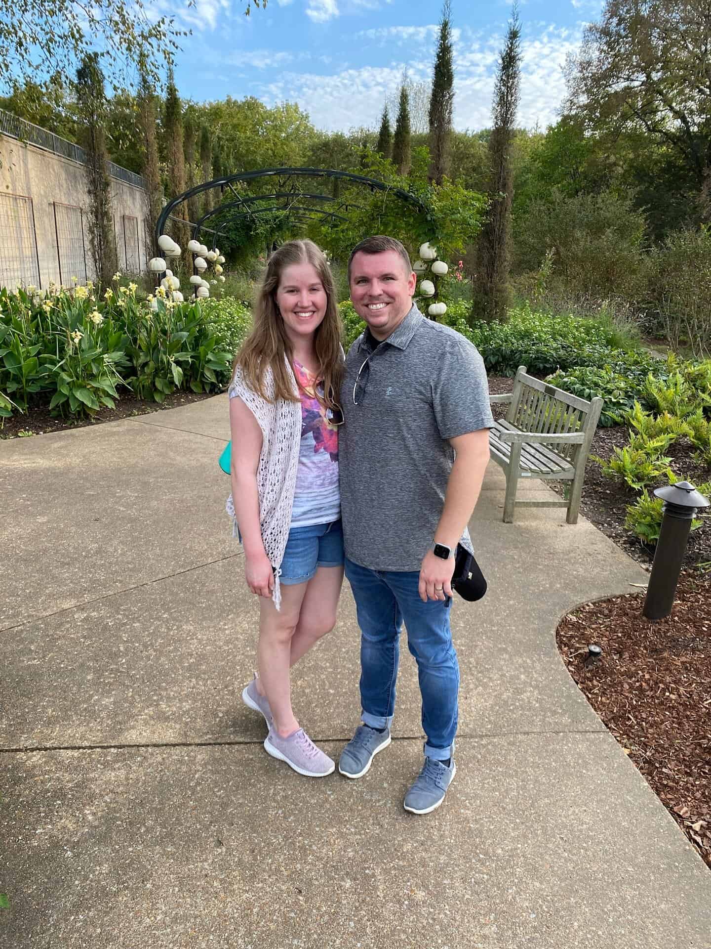 Couple standing on a pathway in a garden with lush greenery and a bench in the background.