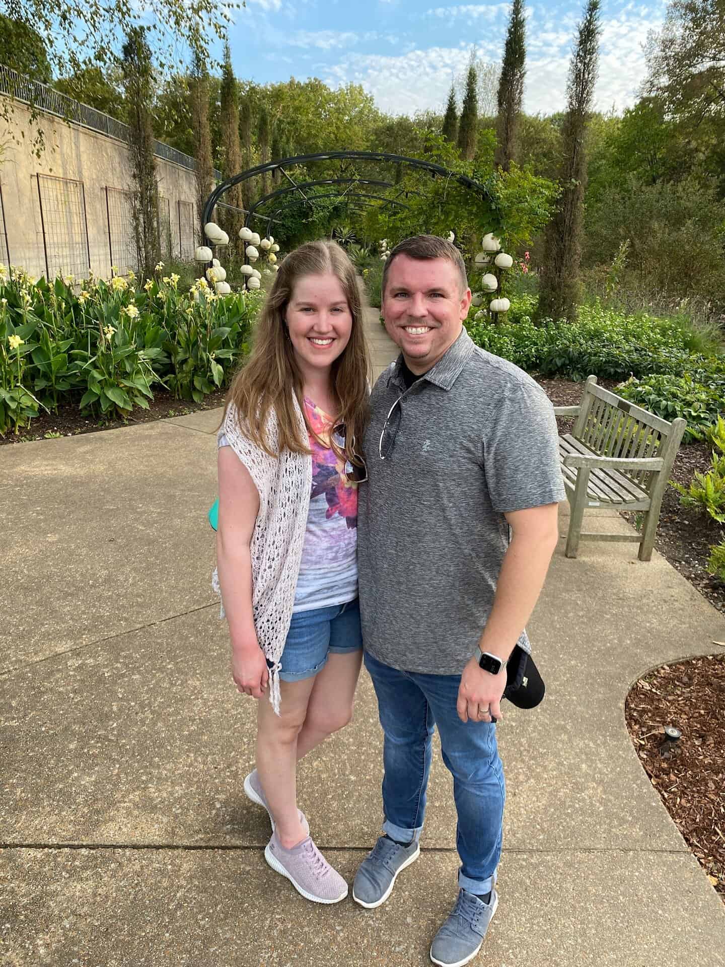 A smiling man and woman stand together on a garden pathway with plants and flowers in the background.