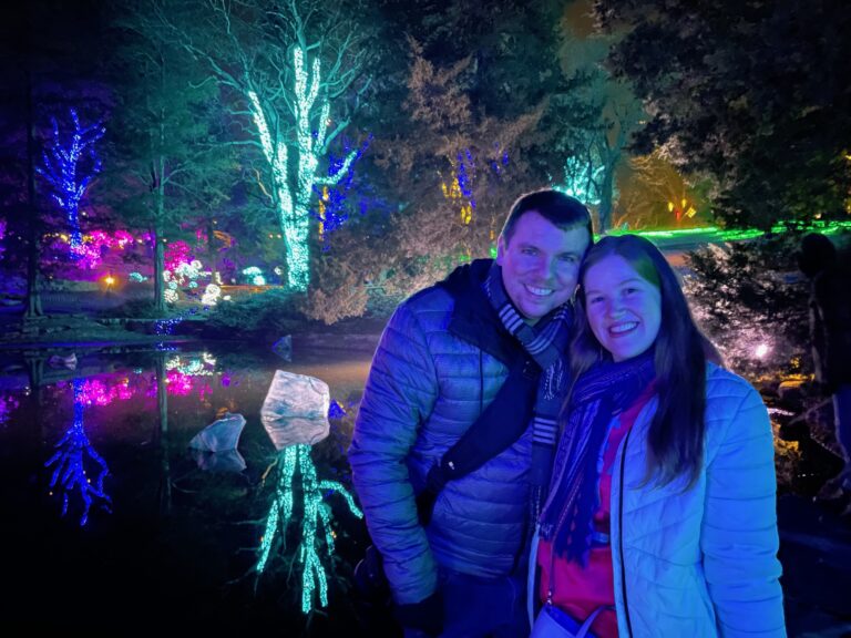 Two people in winter jackets stand smiling in front of a Nashville pond with trees decorated with colorful, illuminated lights at night.