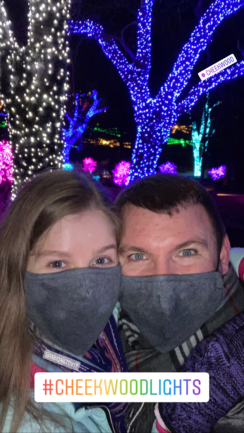Two people wearing gray masks pose for a selfie in front of trees decorated with colorful holiday lights at night. #CHEEKWOODLIGHTS.