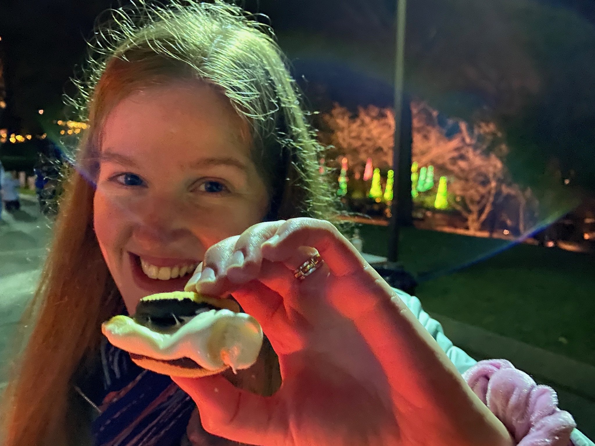 A person smiling and holding a s'more at night in Nashville, with illuminated trees and lights glowing in the blurred background.