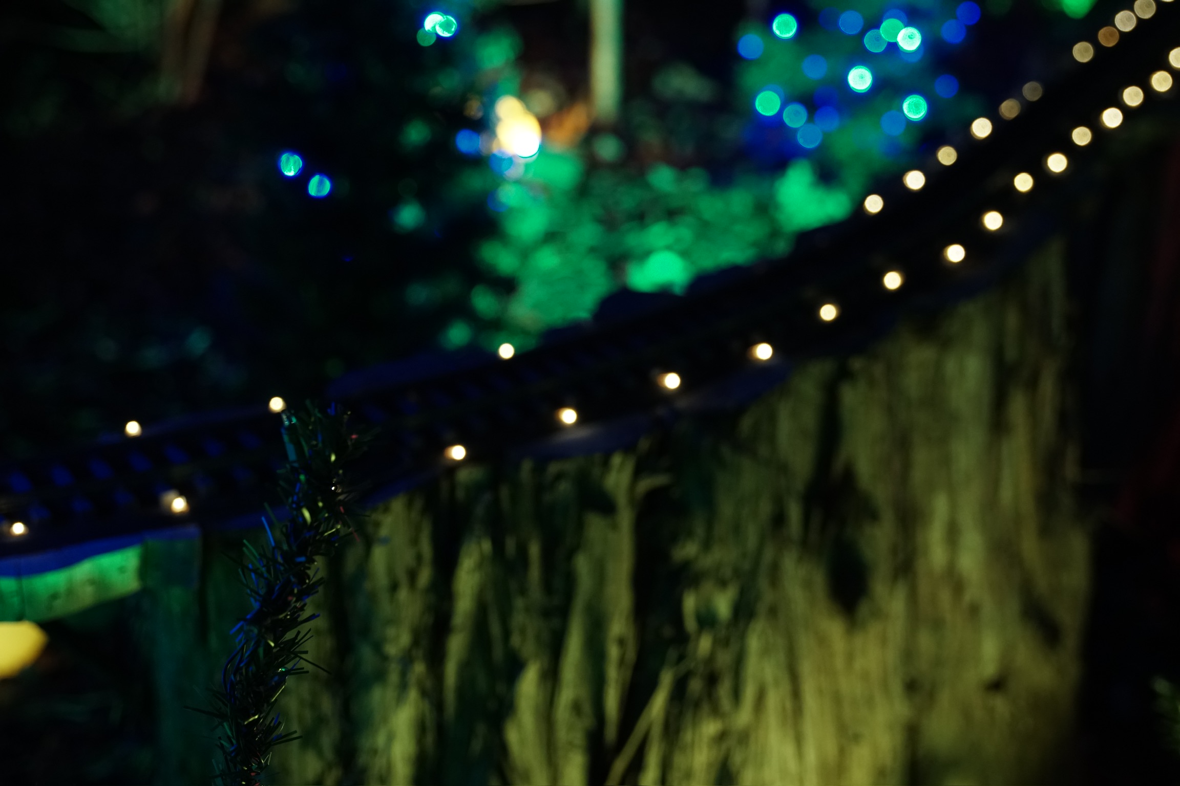 Close-up of a miniature railway track decorated with small lights, set against a blurred background of green foliage and blue lights at night.