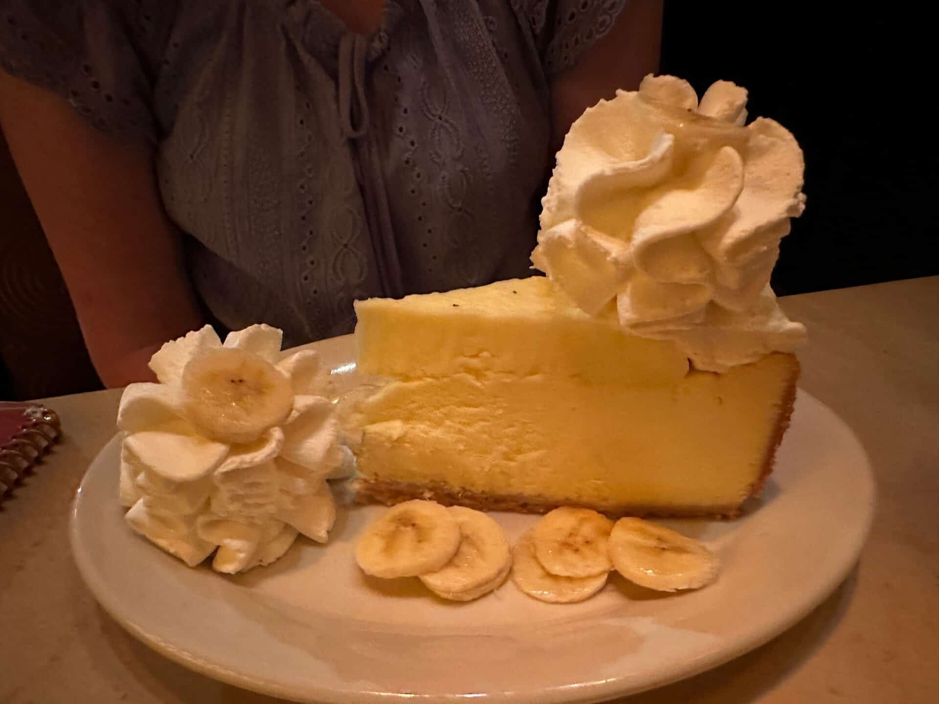 A slice of cheesecake topped with whipped cream and surrounded by banana slices awaits on a pristine white plate, bringing a sweet touch of Nashville charm to your dessert.