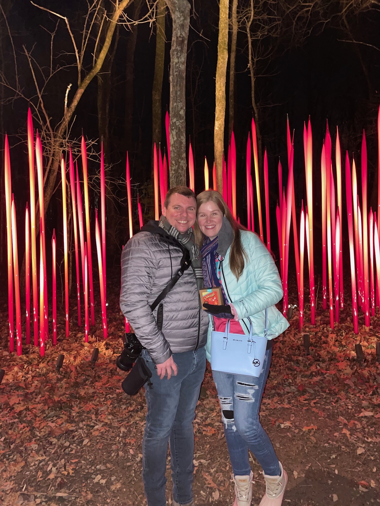 Two people stand close together, smiling, in front of tall red and white glass sculptures in a wooded outdoor setting at night.