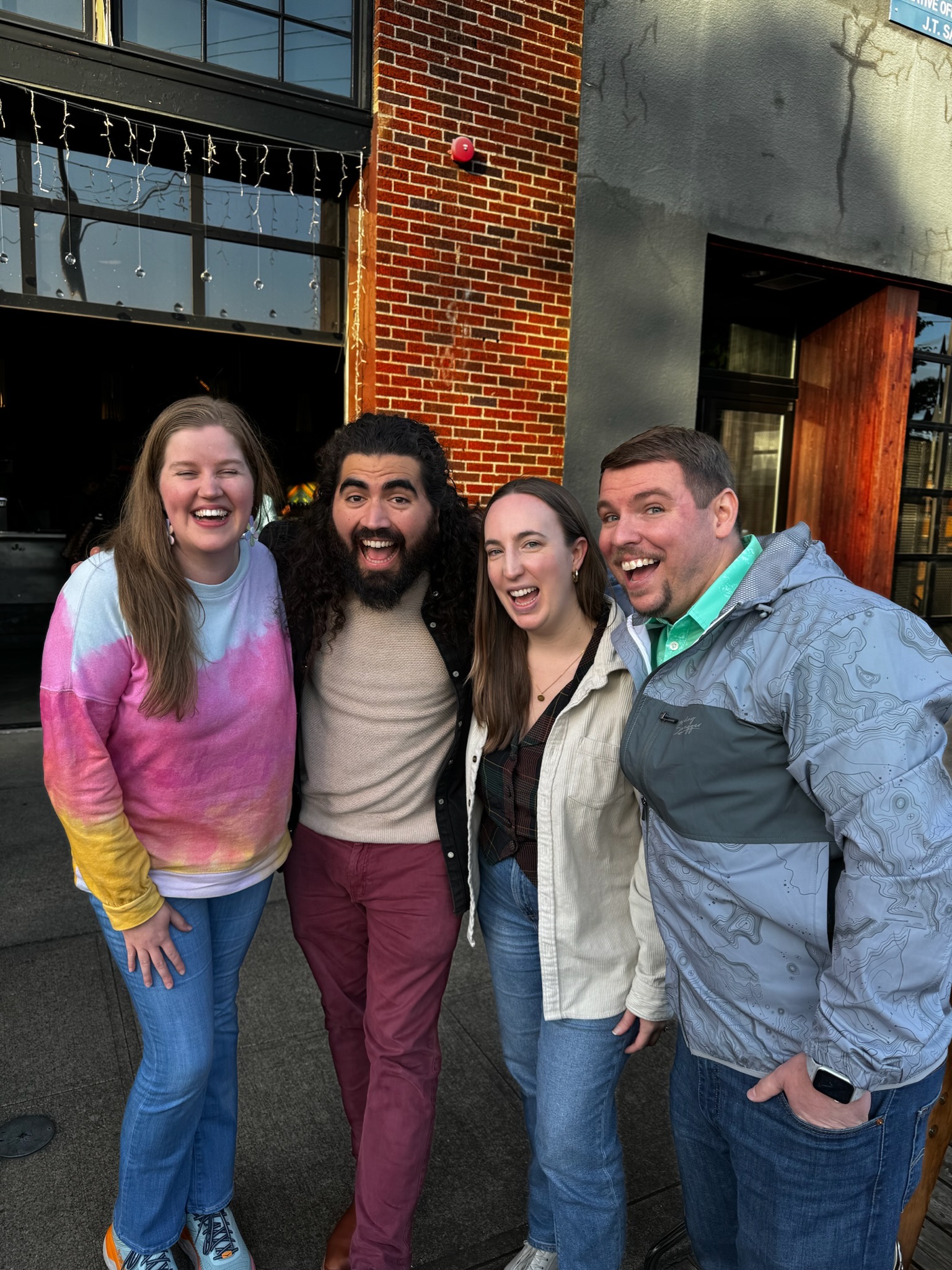 Four adults stand close together outside a brick building, smiling and posing for the camera on a sunny day.