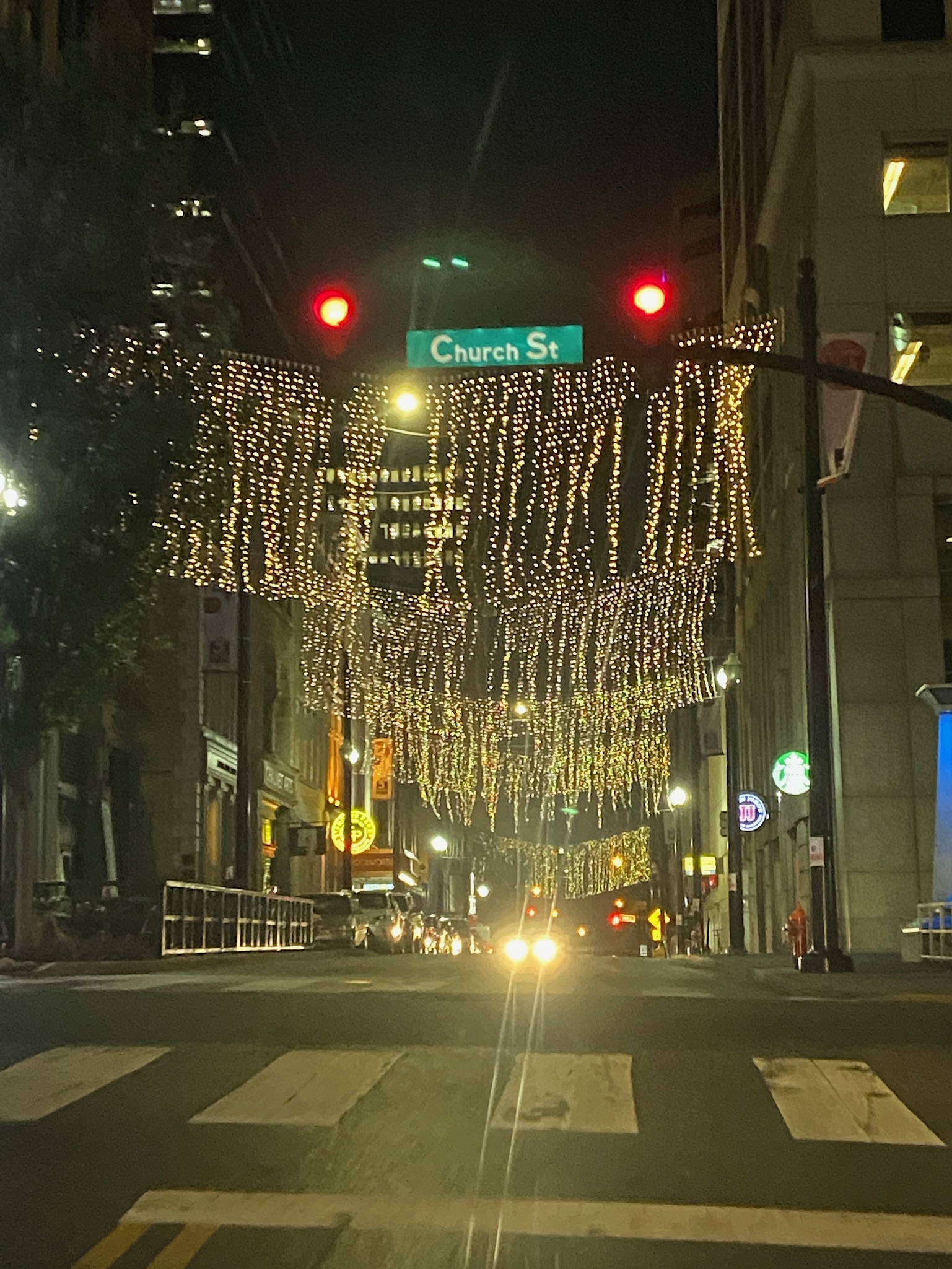 Strings of hanging lights illuminate a city street at night beneath a traffic light and a Church St street sign, with buildings and headlights visible in the background.