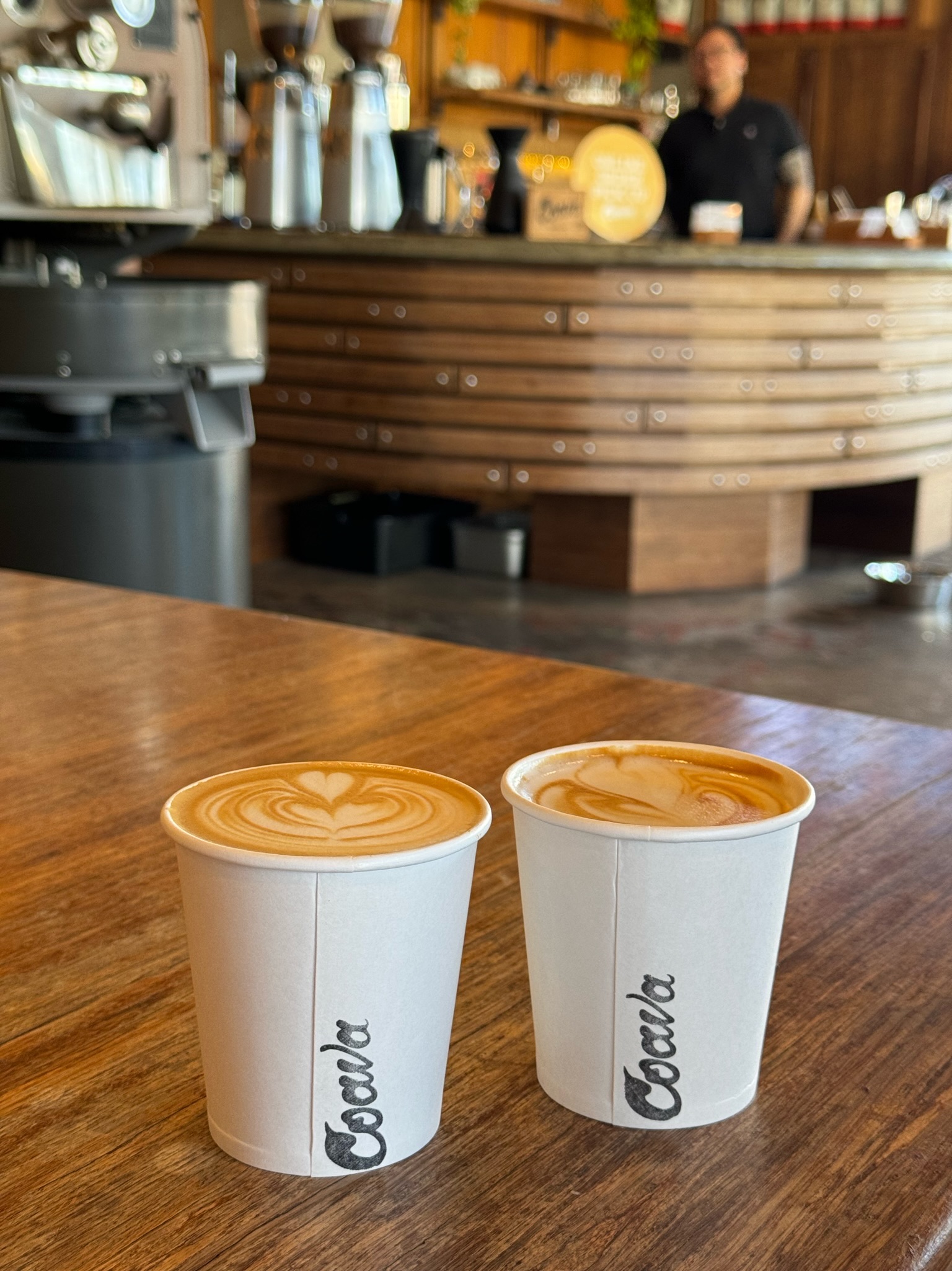 Two paper cups of latte with latte art sit on a wooden table in a coffee shop, with the counter and a barista visible in the background.