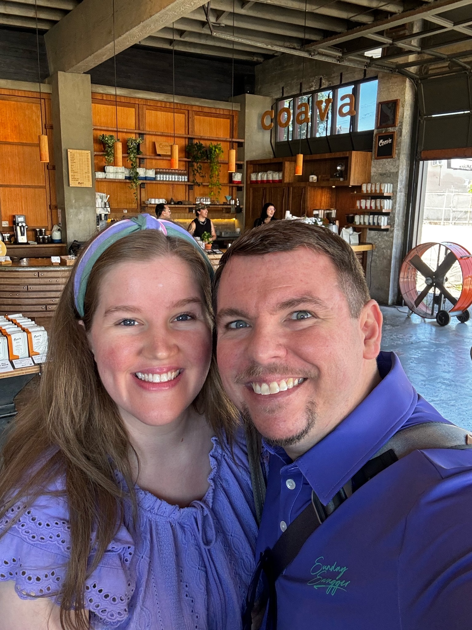 A smiling man and woman pose for a selfie inside a modern coffee shop with wooden decor and a "dava" sign in the background.