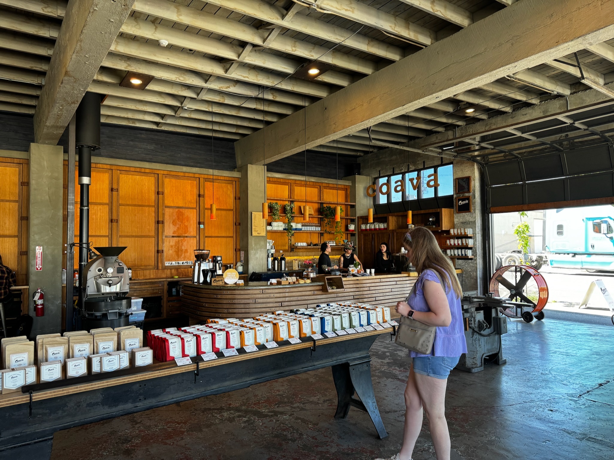 A woman stands in front of a long display of coffee bags inside a modern coffee shop with wood and industrial decor.