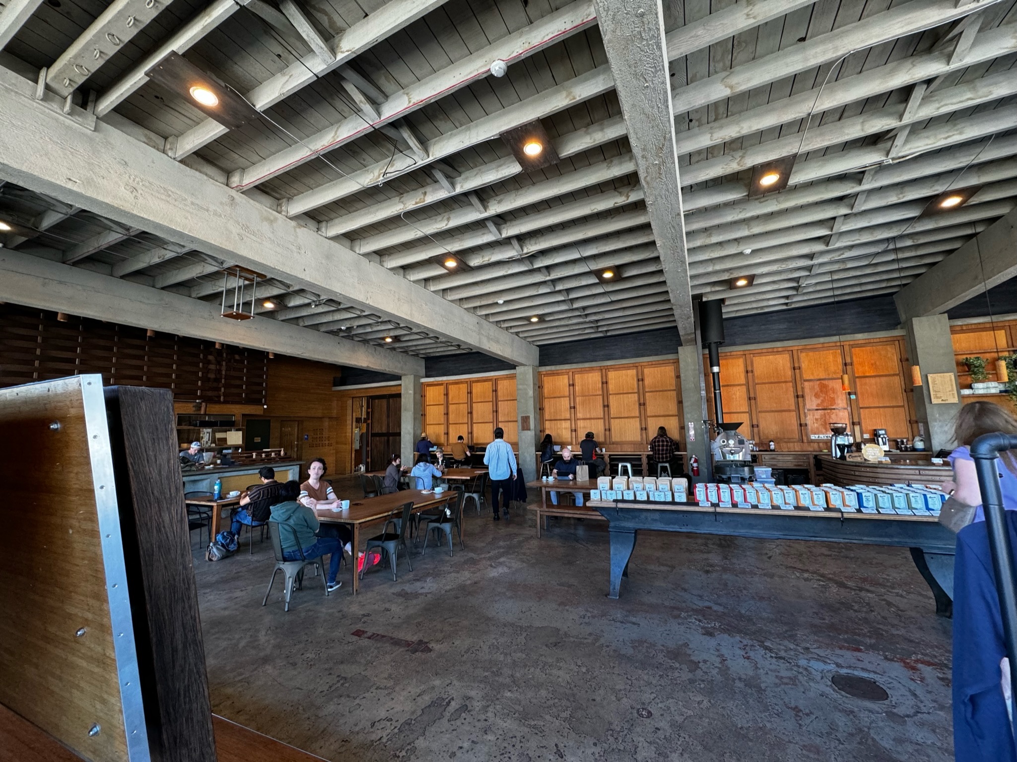 Spacious coffee shop with industrial decor, wooden tables, people sitting and working, and a long counter displaying coffee bags.