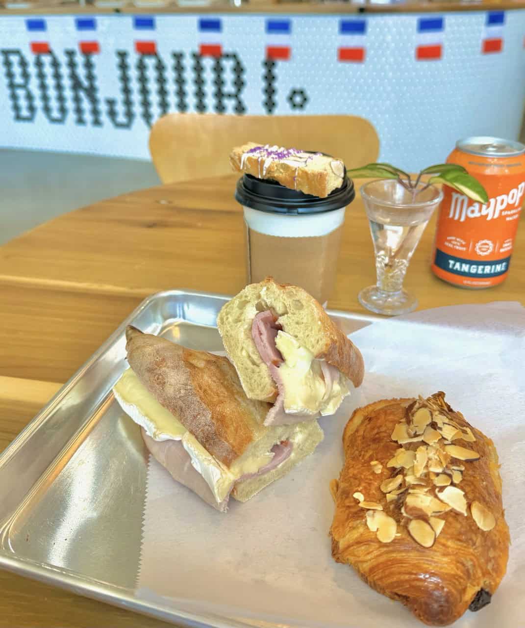 A coffee, tangerine soda, croissant, and ham and cheese baguette sandwich sit next to a vase with a flower on the table. The background features "Bonjour!" in French flag colors, giving this Nashville café moment a delightful European twist.