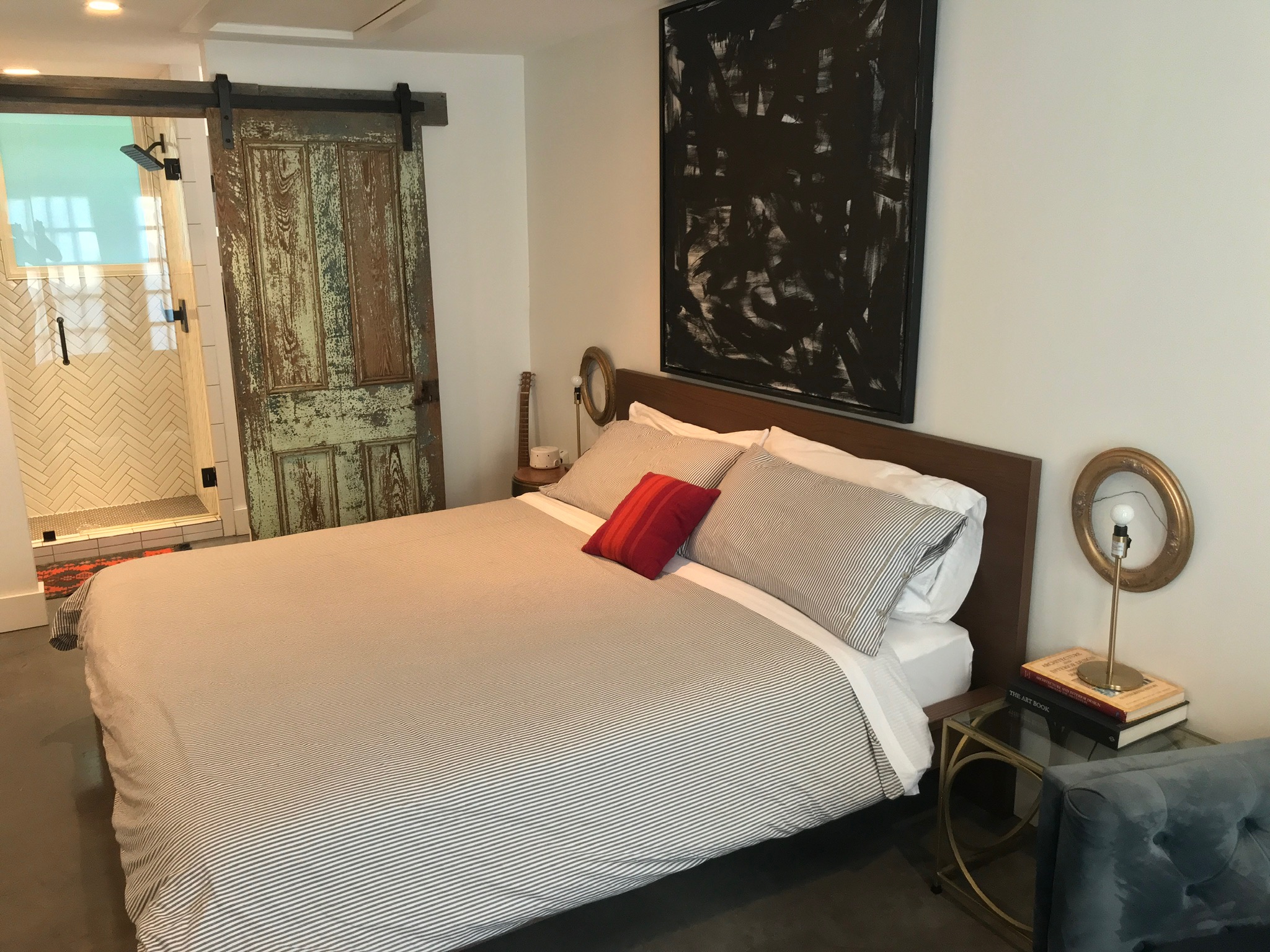 A bedroom with a neatly made bed, gray and white bedding, a single red pillow, bedside tables with lamps, rustic sliding barn door, and abstract art on the wall.