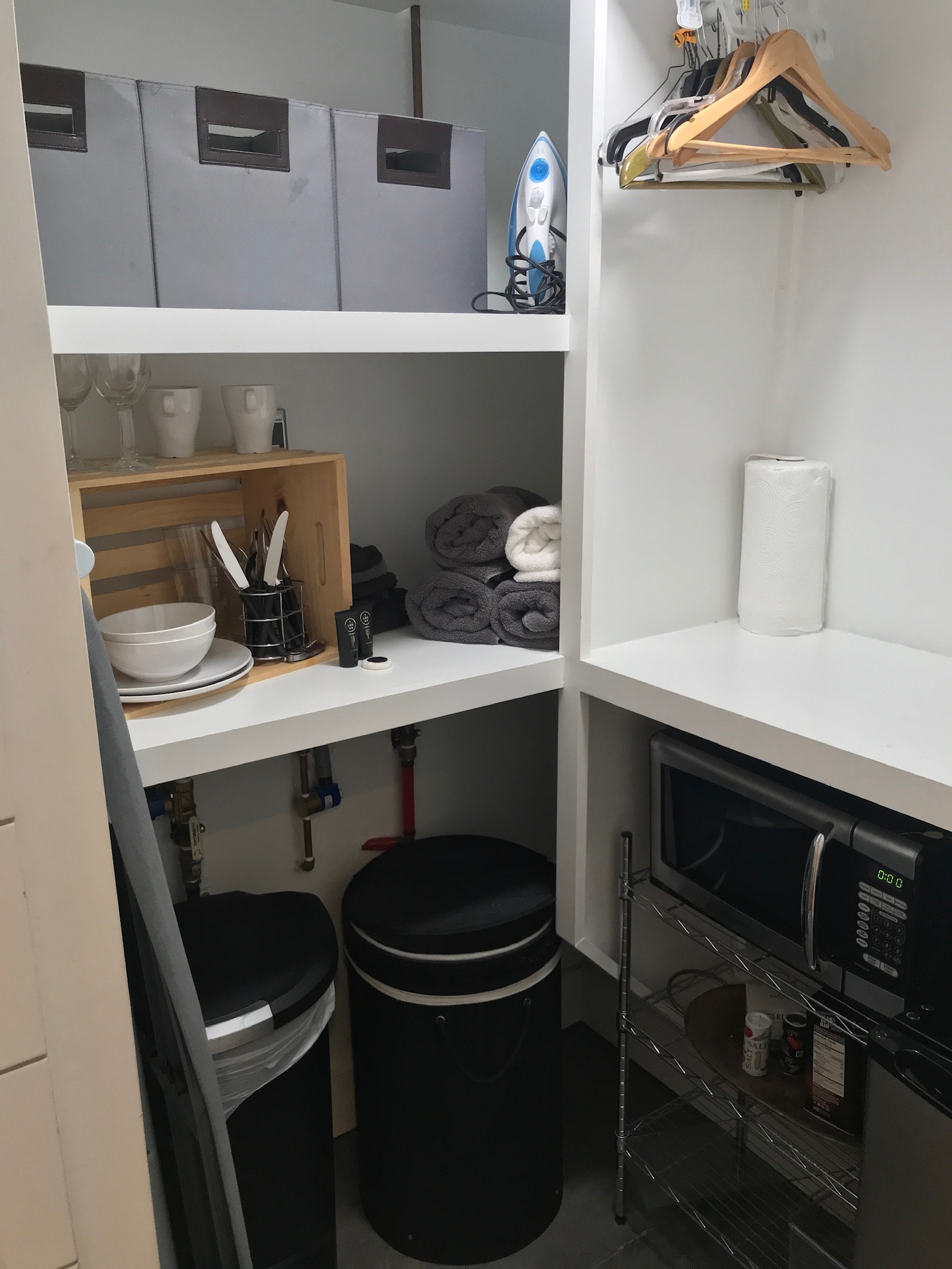 A small storage room with shelves holding boxes, an iron, dishes, glasses, utensils, towels, and a hanging rail with hangers; below are two bins and a microwave on a metal rack.