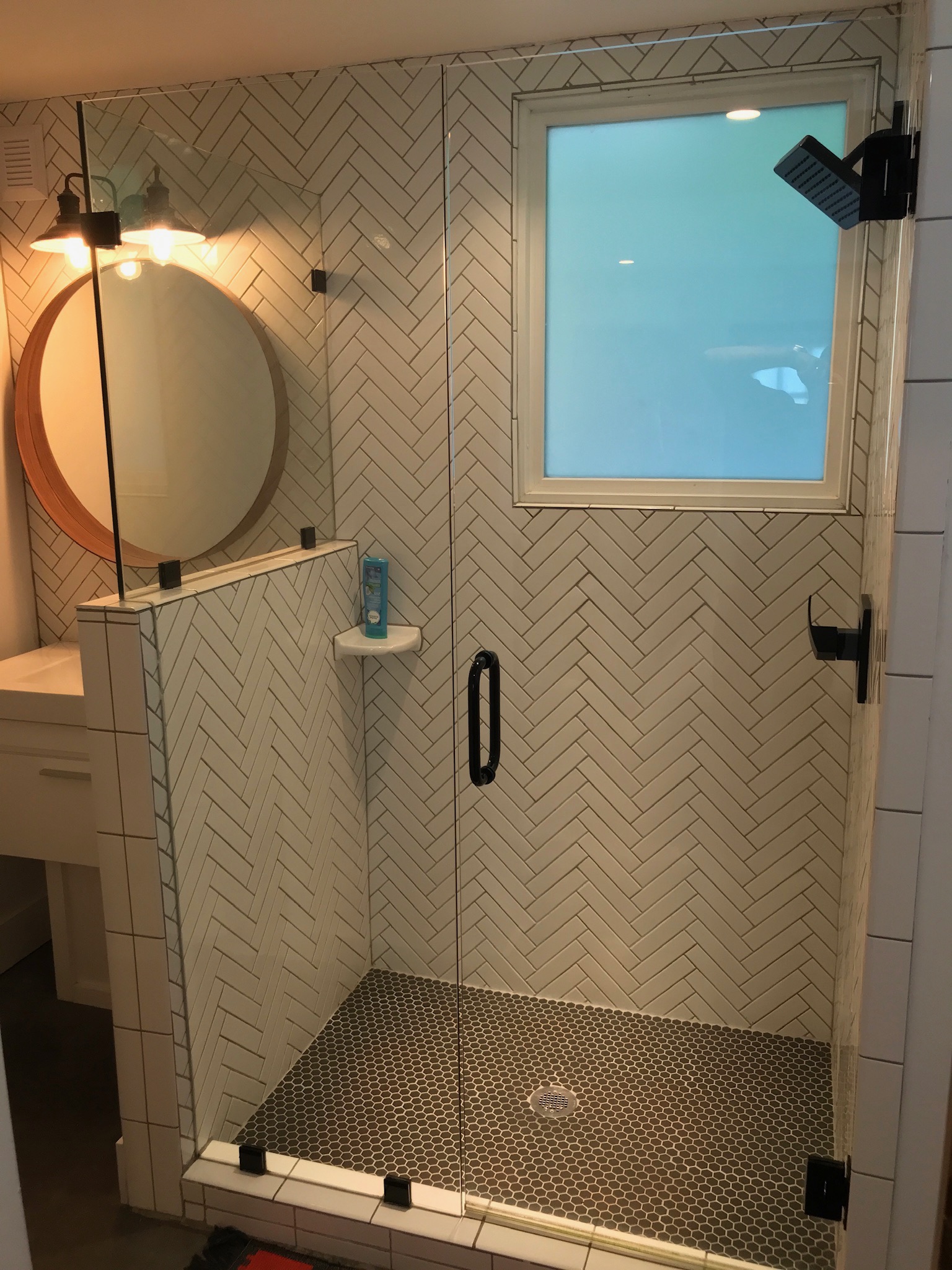 Modern shower with herringbone tile walls, black fixtures, a glass door, a round mirror, and a small window above the shower area. A bottle is placed on a built-in corner shelf.
