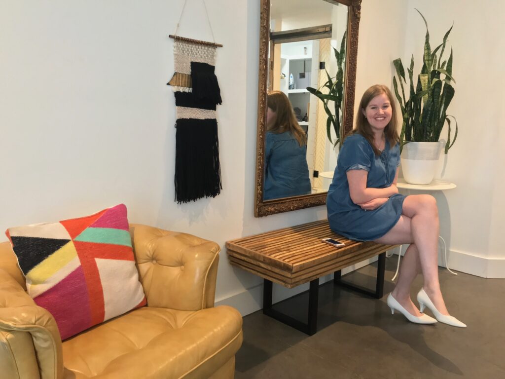 Woman in a blue dress sits on a wooden bench next to a large mirror, with a plant and a yellow armchair nearby in a modern room.