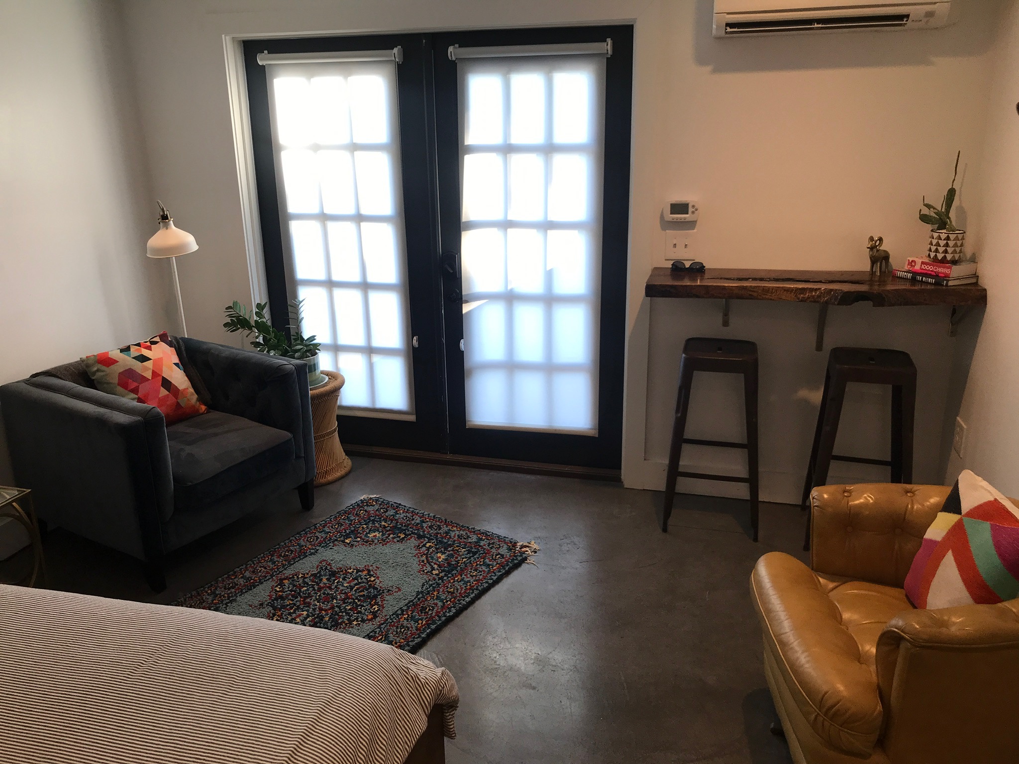 A modern room with a bed, two armchairs, a small rug, a tall counter with two stools, and frosted glass double doors letting in light.