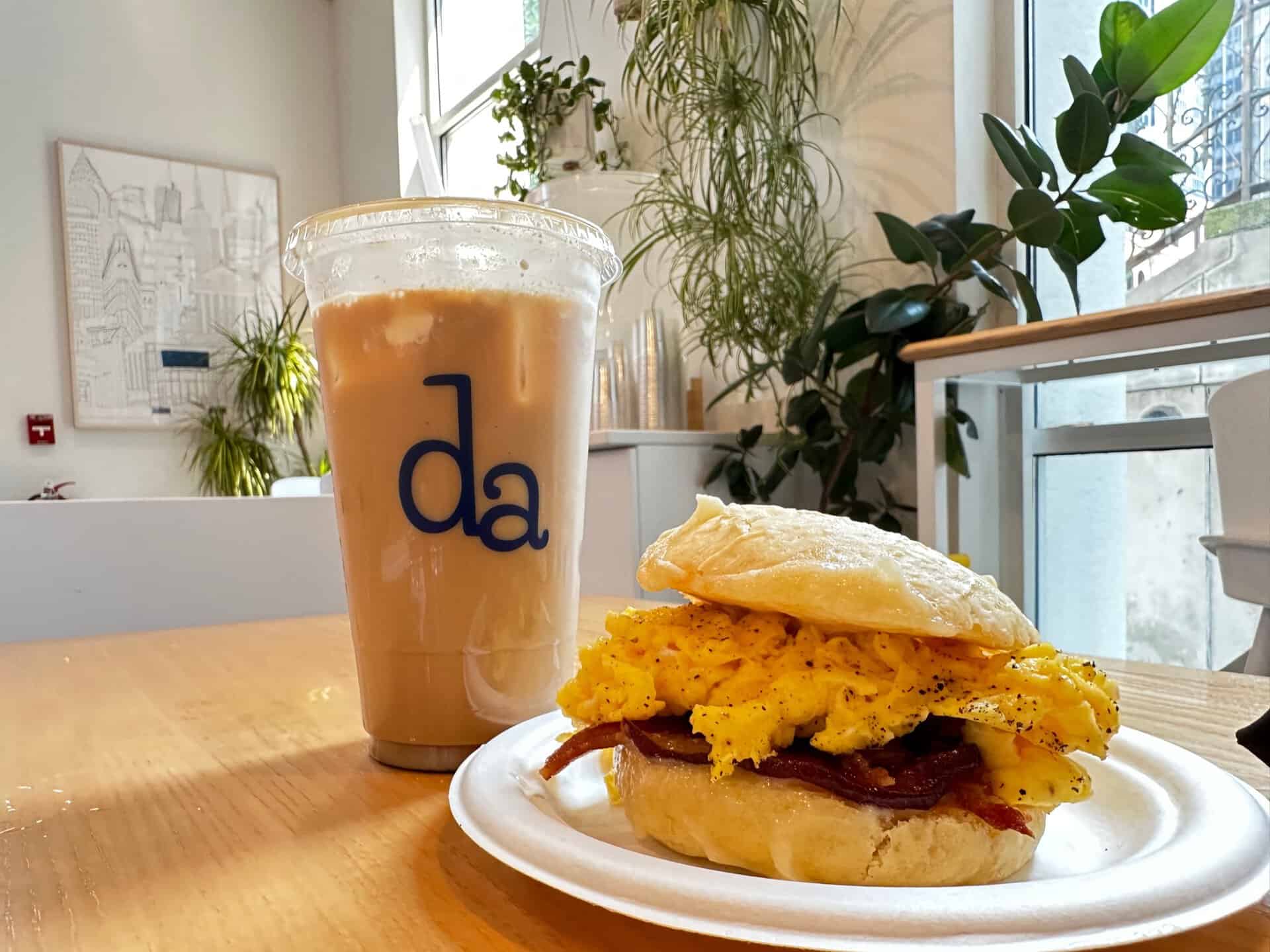 Iced coffee with "da" logo and a Nashville-inspired breakfast sandwich featuring scrambled eggs and bacon on a plate. Indoor setting with plants in the background.
