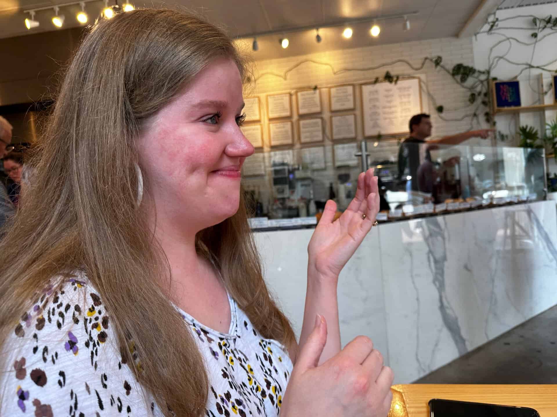 A woman smiles warmly, gesturing with her hands as she relaxes in a Nashville café, surrounded by a marble counter and lush plants.