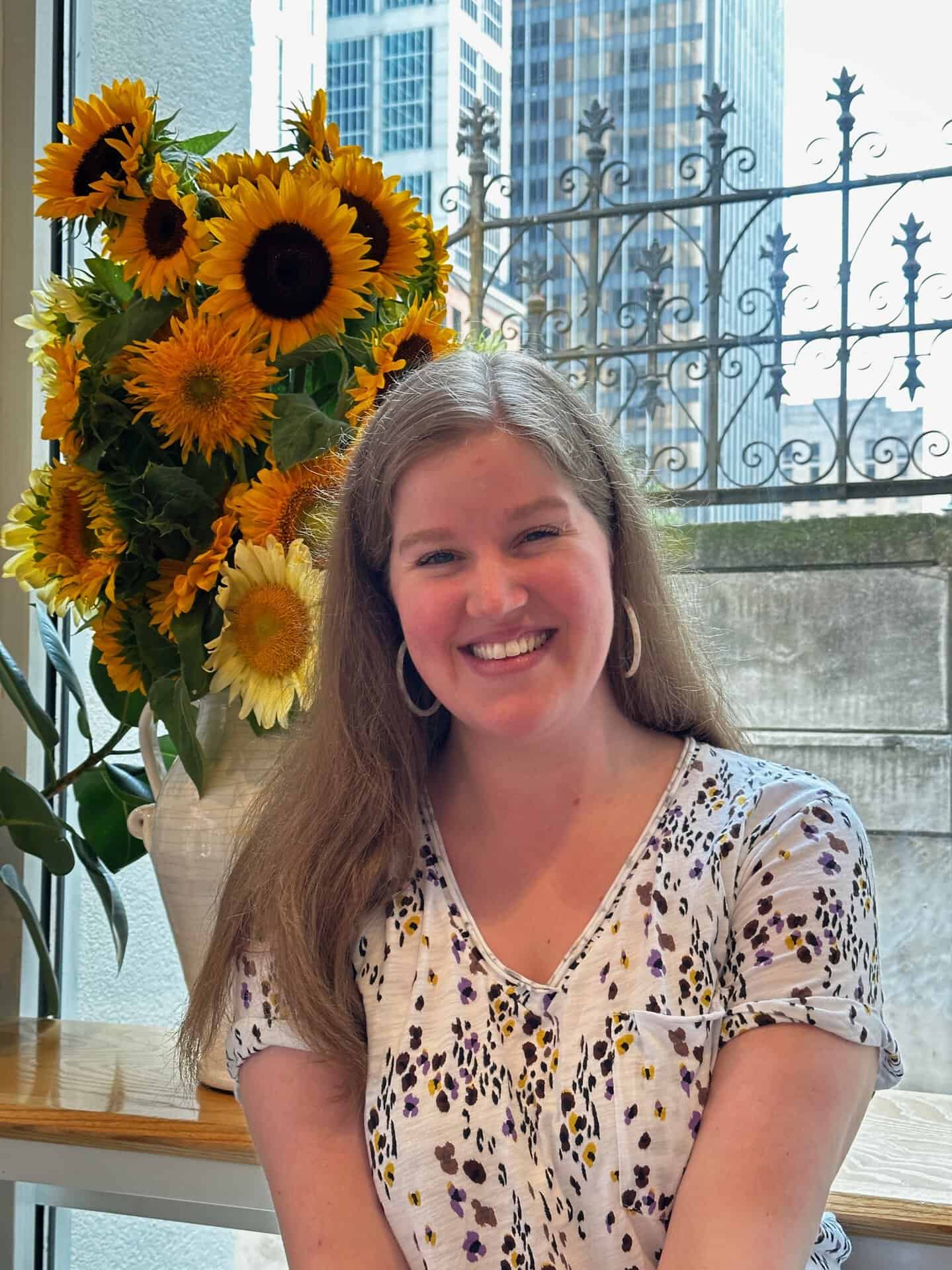 A woman with long hair smiles warmly in front of a vase of sunflowers, her patterned top adding flair. Through the window, the vibrant cityscape of Nashville's tall buildings creates a striking backdrop.