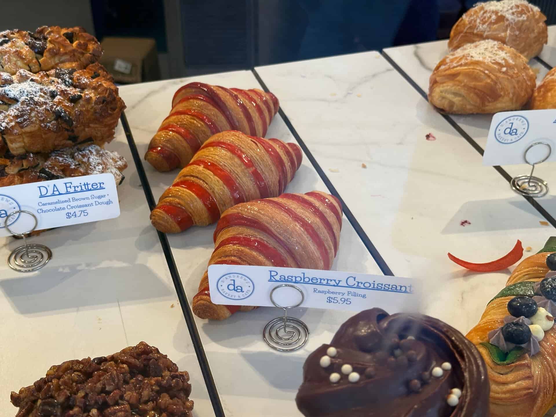 A delightful selection of pastries graces the Nashville counter, featuring raspberry croissants at $5.95 alongside the famous DA Fritter for $4.75. Clear signs display each tempting treat's name and price.