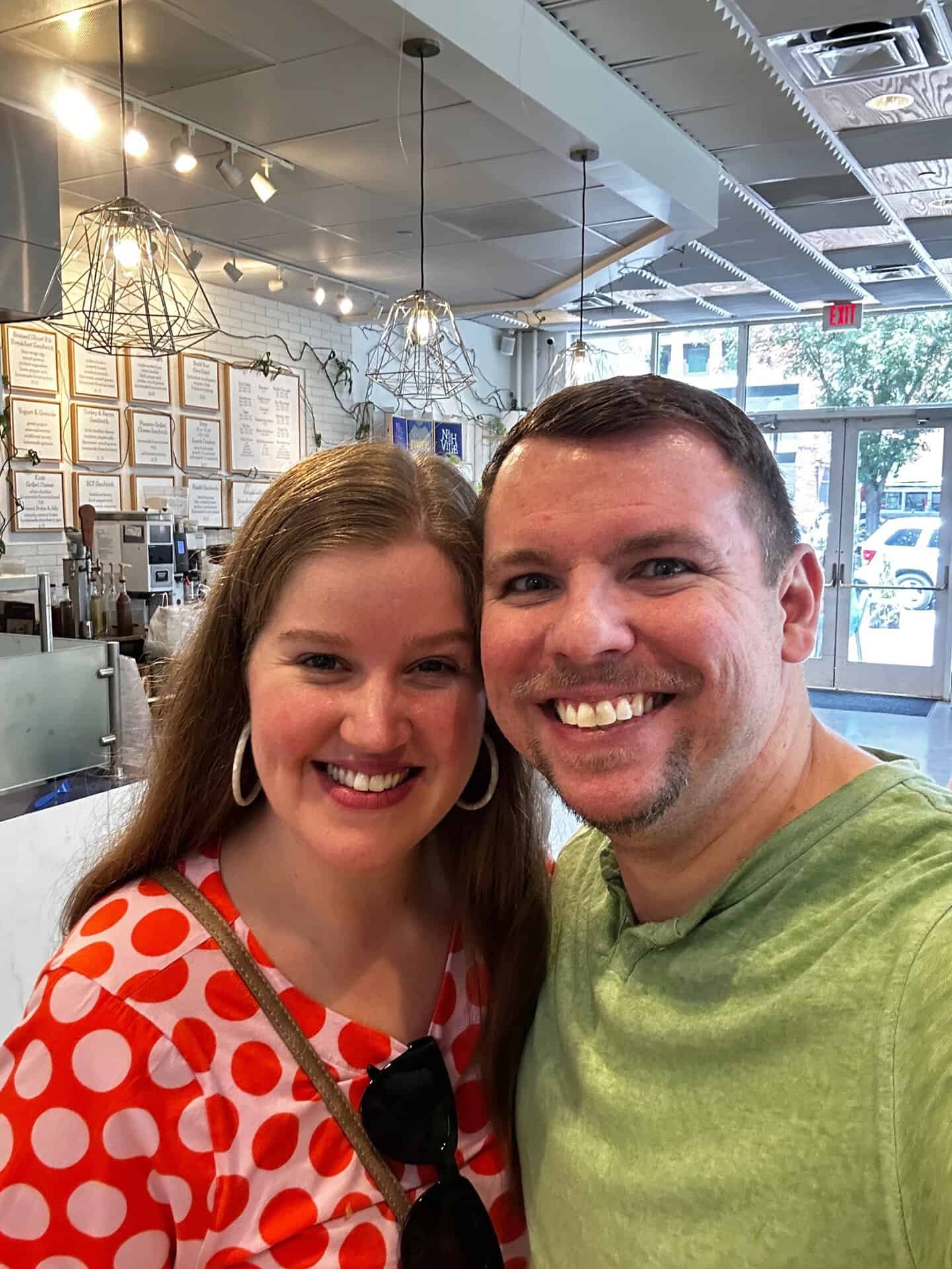 A smiling man and woman snap a selfie inside a modern Nashville cafe, with the menu visible in the background. She sports a red polka dot top, while he dons a green shirt, capturing their fun outing in Music City.