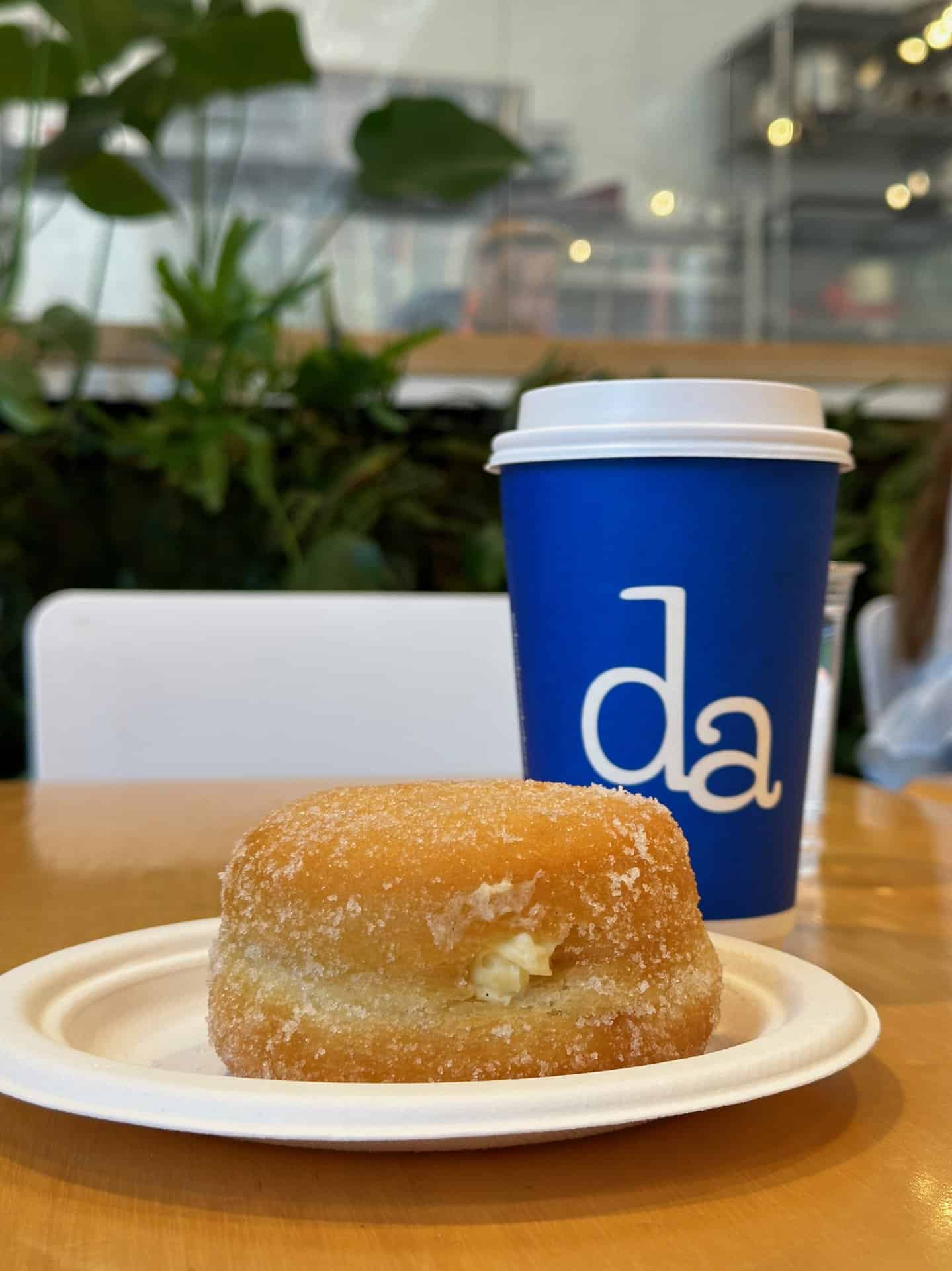 A sugar-coated doughnut rests on a plate alongside a blue coffee cup adorned with "da" letters, all set on a rustic wooden table indoors. This cozy Nashville setting invites you to savor every bite and sip.