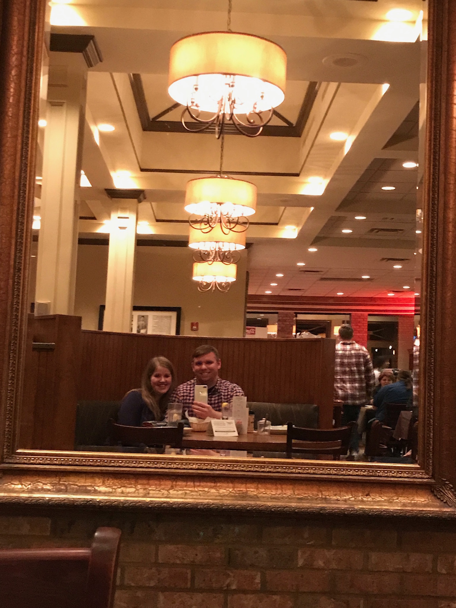 A couple sits at a table in a restaurant, their reflection captured in a large framed mirror with hanging chandeliers visible above.