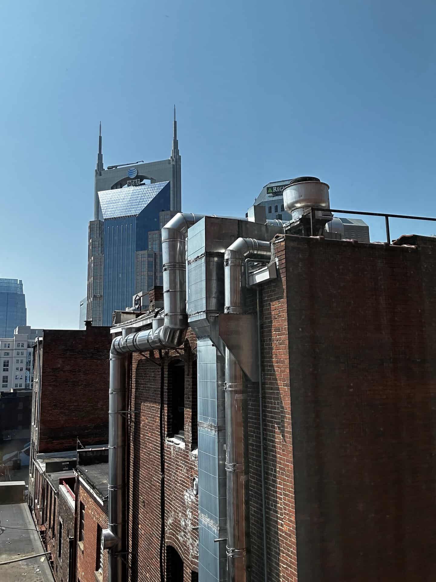 Rooftop view of an urban area with brick buildings and multiple exposed pipes, reminiscent of Nashville's skyline. A tall, pointed skyscraper stands proudly against the clear blue sky in the background.