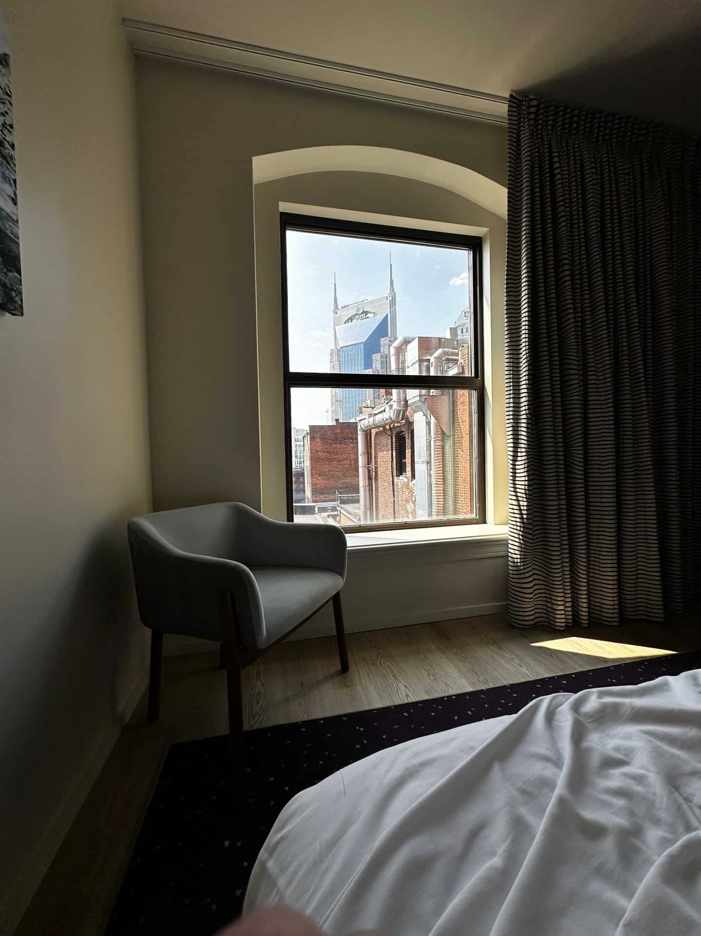 A chair by the window offers a view of a distant building shaped like a tower, reminiscent of Nashville's skyline. Sunlight filters through, casting playful shadows on the curtains and floor.