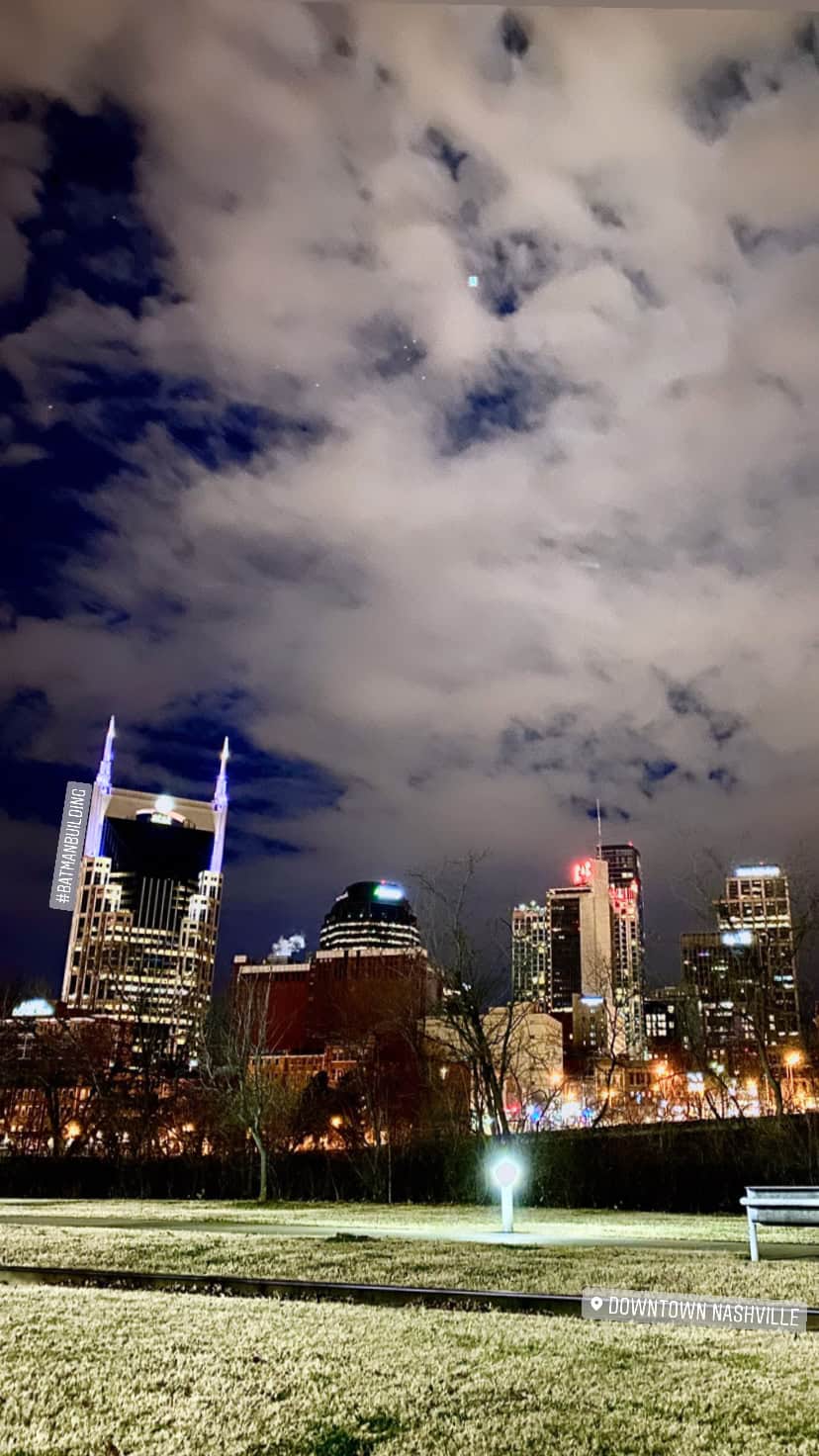 A nighttime view of downtown Nashville’s skyline under a partly cloudy sky, with illuminated buildings and a grassy foreground.
