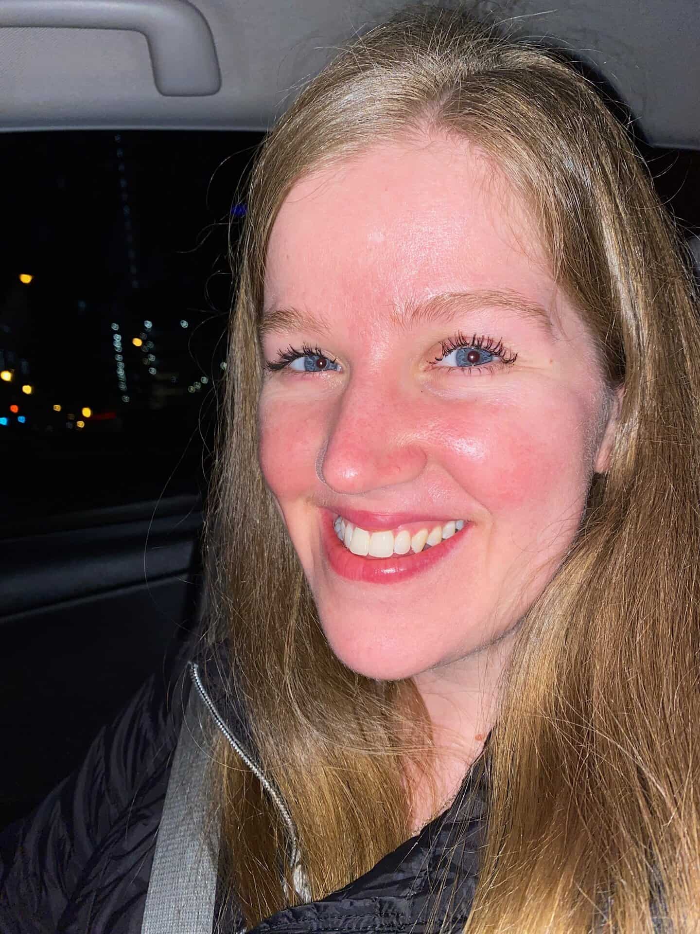A woman with long blonde hair smiles at the camera while sitting in a car at night, wearing a black jacket and a seatbelt.