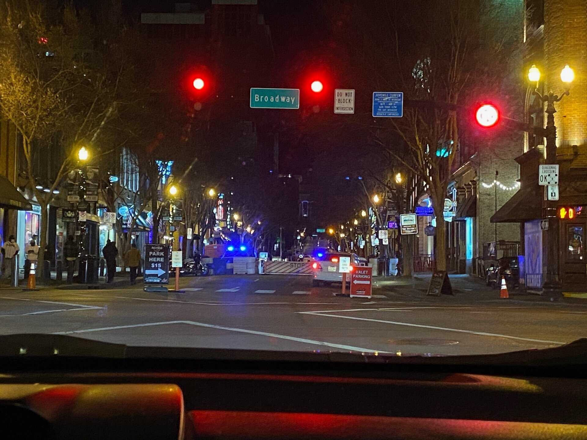 A nighttime street scene shows a road closure with police cars, barricades, and flashing lights on Broadway under a red traffic light. Pedestrians are visible on sidewalks.