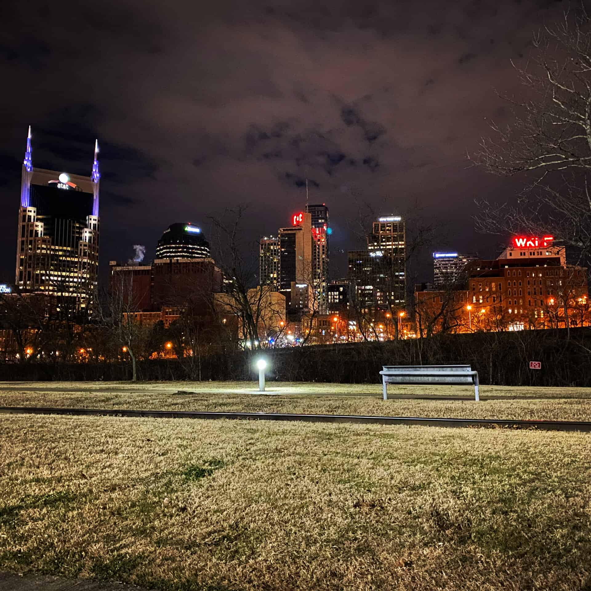 A view of a city skyline at night with illuminated buildings and a grassy park in the foreground featuring a single empty bench.