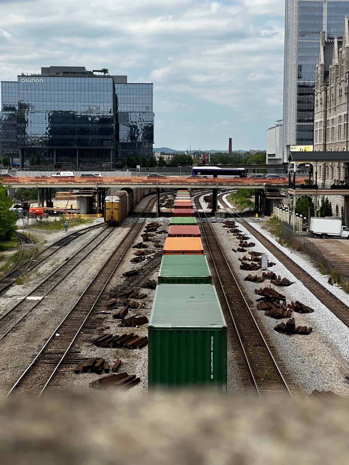 A bustling railway yard in Nashville showcases multicolored cargo containers on tracks, all framed by urban buildings under a cloudy sky.