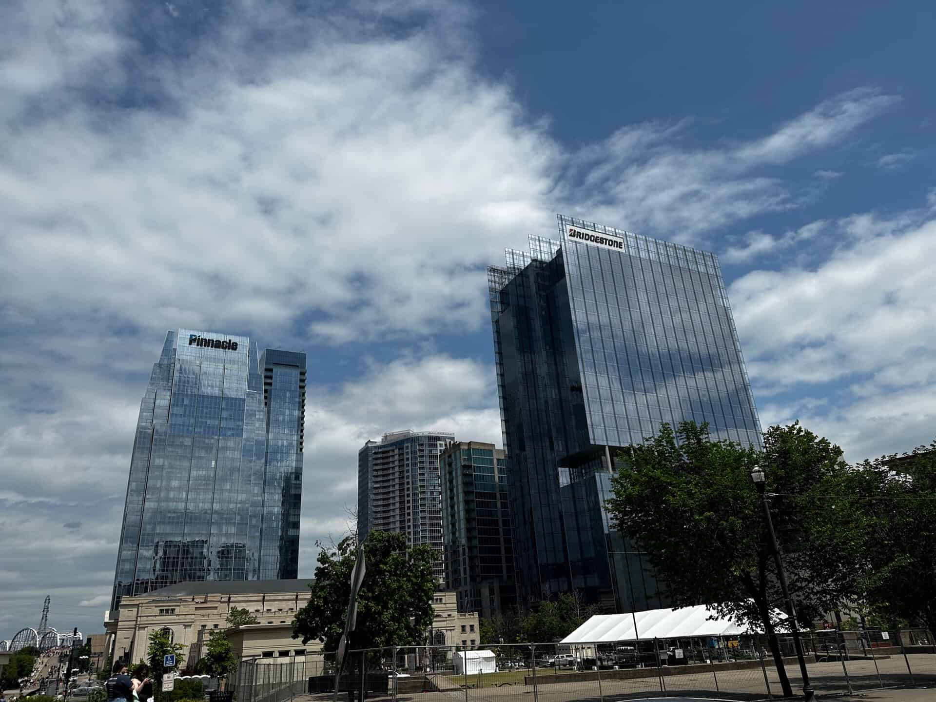 Tall glass skyscrapers under a cloudy Nashville sky, surrounded by trees and a white tent in the foreground.