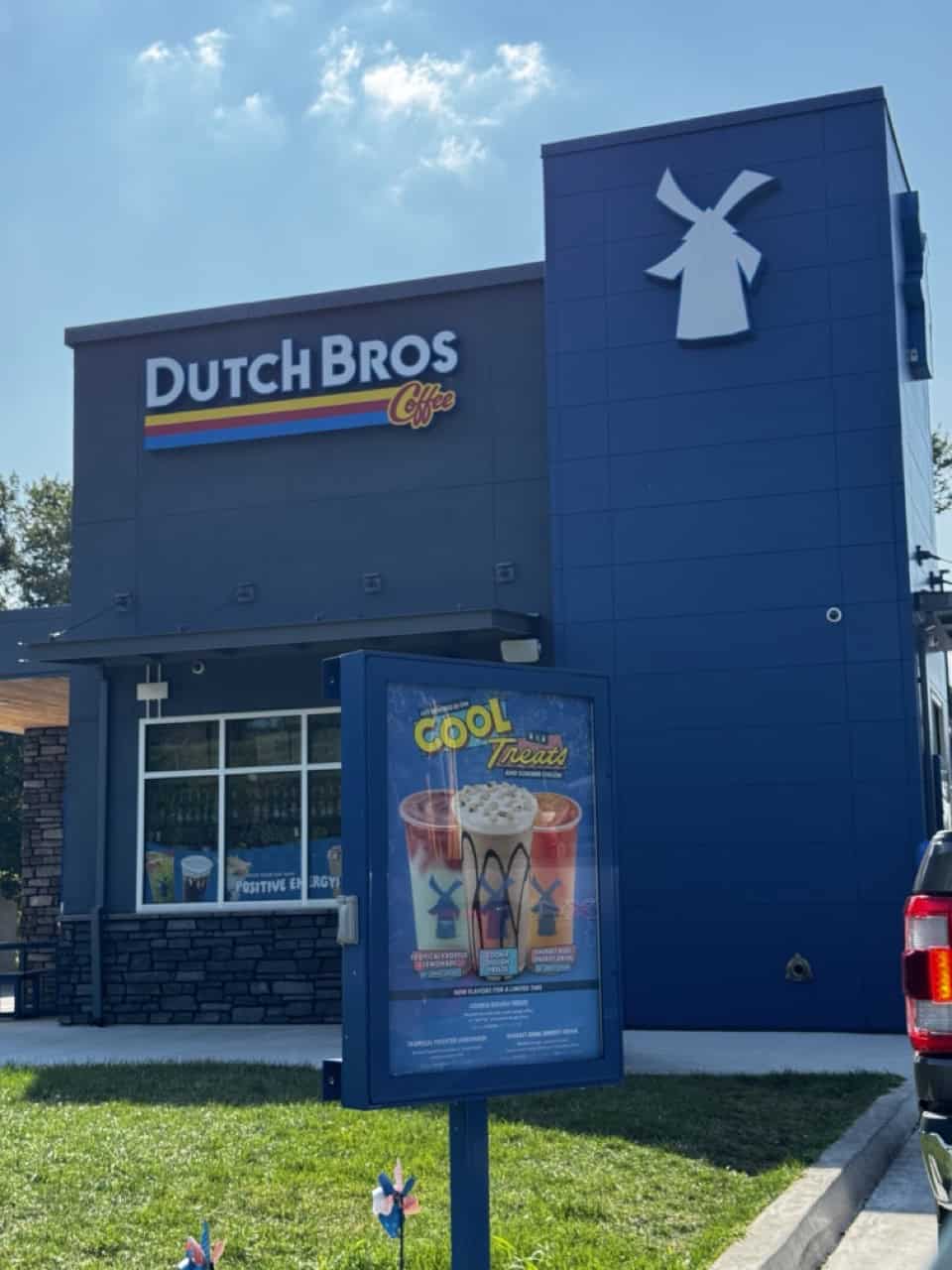 The exterior of a Dutch Bros Coffee location, Nashville charm meets the iconic windmill logo and promotional poster. A vehicle is partially visible on the right, adding to the bustling energy.