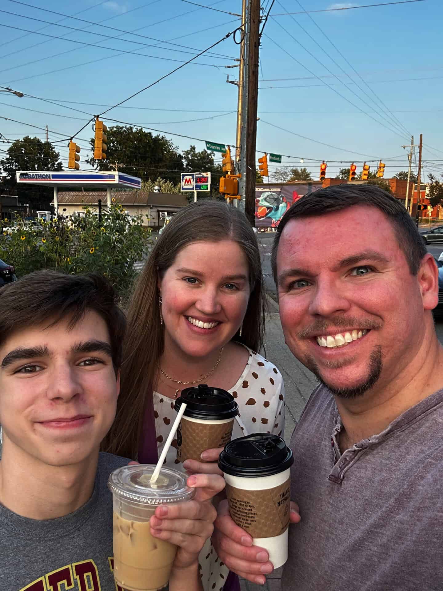 Three people are posing and smiling while holding coffee cups near an intersection in Nashville, with traffic lights and a gas station visible in the background.