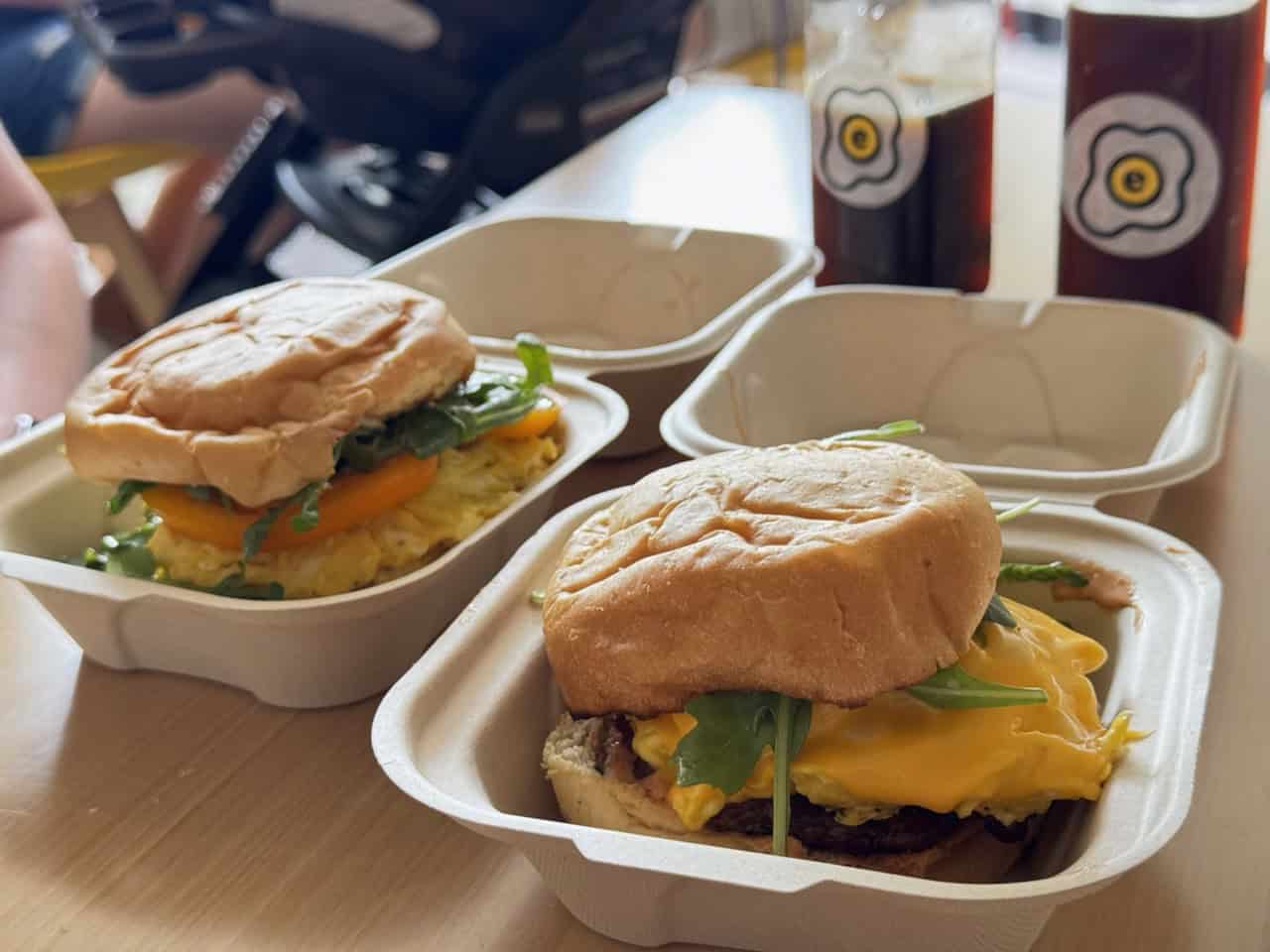 Two Nashville-style burgers with various toppings sit temptingly on buns in white takeout boxes on the table, while two bottles of drink linger invitingly in the background.